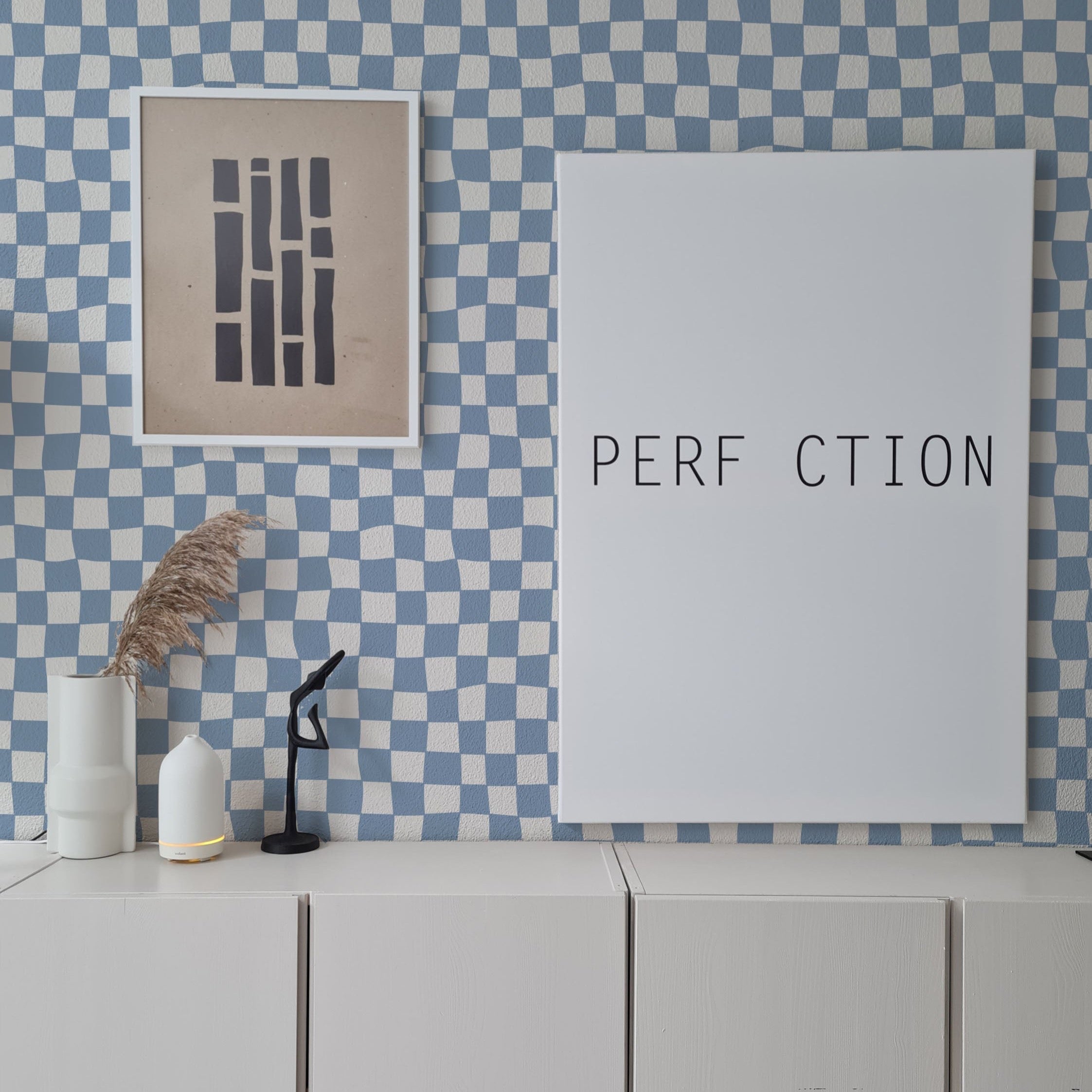 A minimalistic interior design featuring a blue and white checkered wallpaper that covers the entire room. A framed abstract art piece hangs on the left, beside a large poster with the text "PERFECTION" missing the first and last letters. The decor includes a small sculpture, a couple of modern vases, and a cylindrical lamp on a low white cabinet.