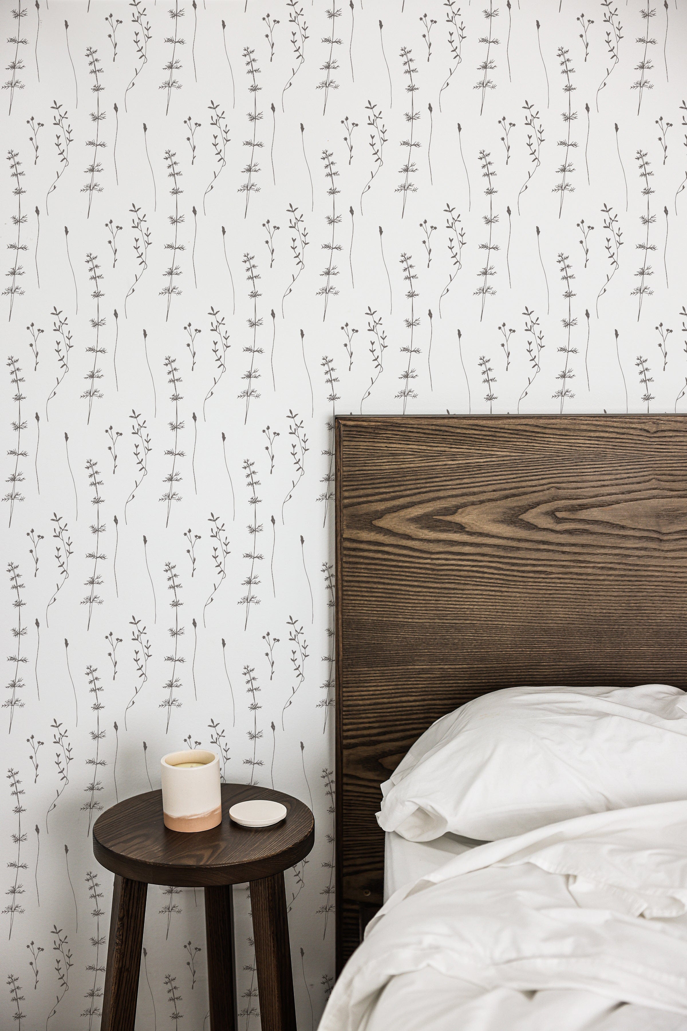 A minimalist bedroom scene featuring Delicate Wildflower Wallpaper. A wooden bed frame adjacent to a small round side table, which holds a ceramic candle holder, enhances the organic and tranquil ambiance created by the floral pattern.