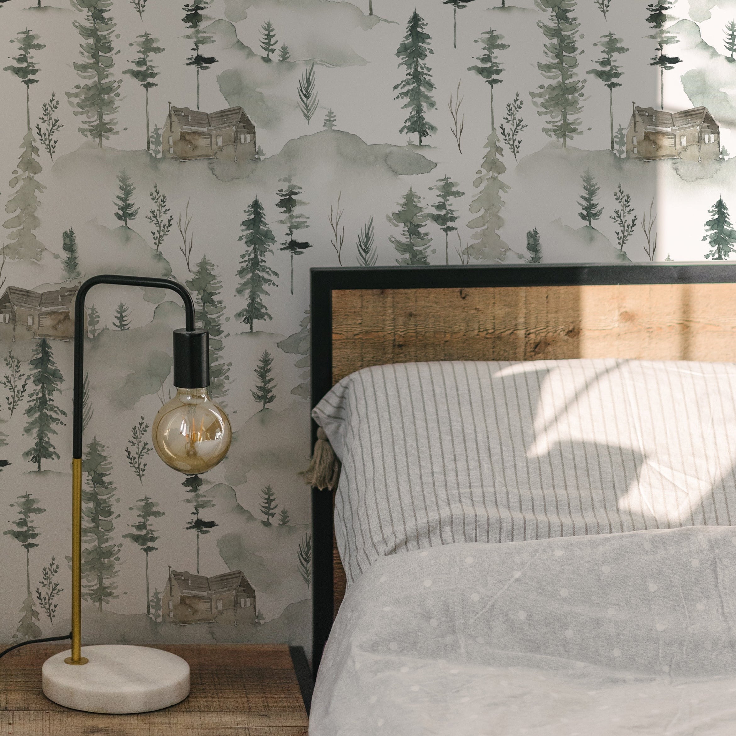 Bedroom interior showcasing In the Woods Wallpaper, creating a peaceful backdrop with its delicate forest and cabin motifs. The room includes a simple bed with striped bedding and a stylish bedside lamp, complementing the natural theme