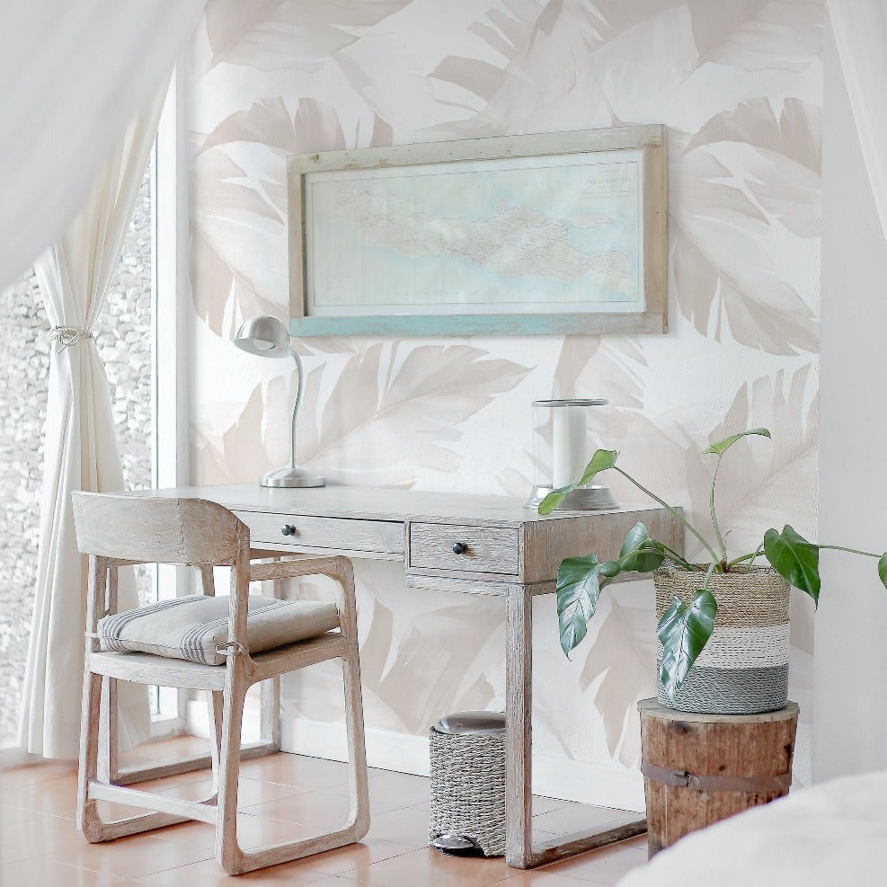 The neutral tropical wallpaper is displayed in an elegant home office setting, offering a backdrop to a vintage-inspired wooden desk and chair. The room's decor, complete with light drapery, a framed map, and potted plants, exudes a sense of calm and sophistication.