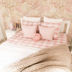 A cozy bedroom scene with a white bed dressed in pink and white checkered bedding and pastel pillows. The background features Floral Tropical Wallpaper, creating a serene and inviting atmosphere with its soft tropical foliage design.