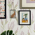 A stylish wall decorated with Lavender Watercolour Wallpaper, adorned with various framed art pieces. The wallpaper’s delicate lavender and green leaf patterns create a serene and artistic backdrop.