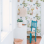 A bright and airy room with the Watercolour Bouquet Floral Wallpaper. The wallpaper has a soft floral design in pastel hues, enhancing the fresh and tranquil ambiance. The space includes a white desk with a plant and a decorative lamp, and a blue chair, adding a pop of color against the floral backdrop.