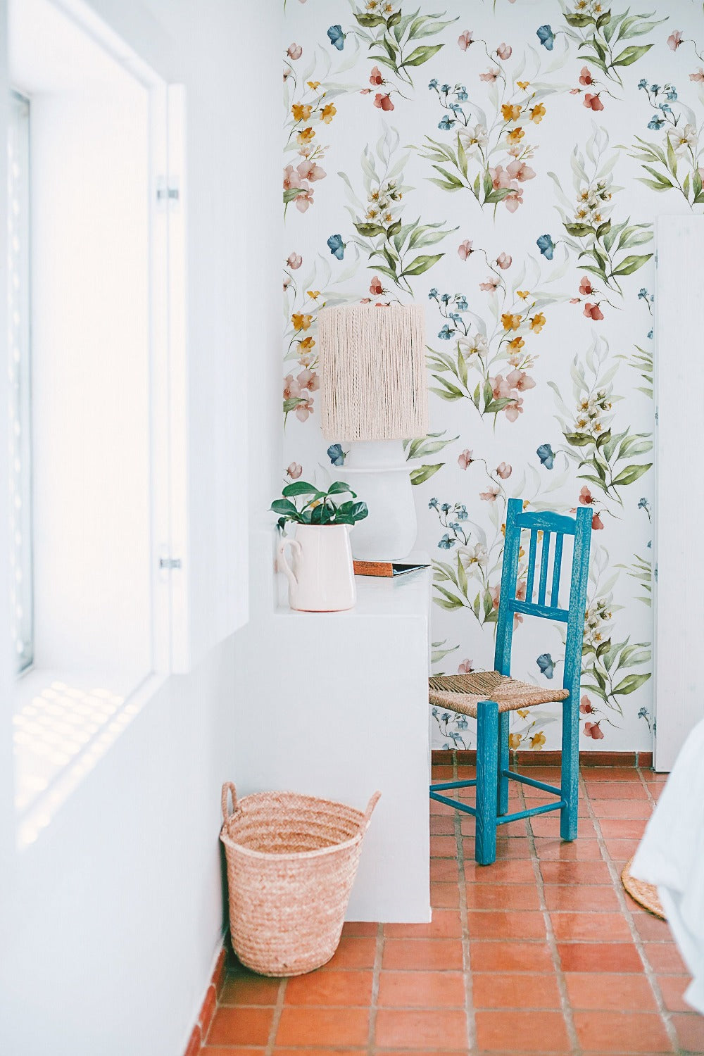 A bright and airy room with the Watercolour Bouquet Floral Wallpaper. The wallpaper has a soft floral design in pastel hues, enhancing the fresh and tranquil ambiance. The space includes a white desk with a plant and a decorative lamp, and a blue chair, adding a pop of color against the floral backdrop.