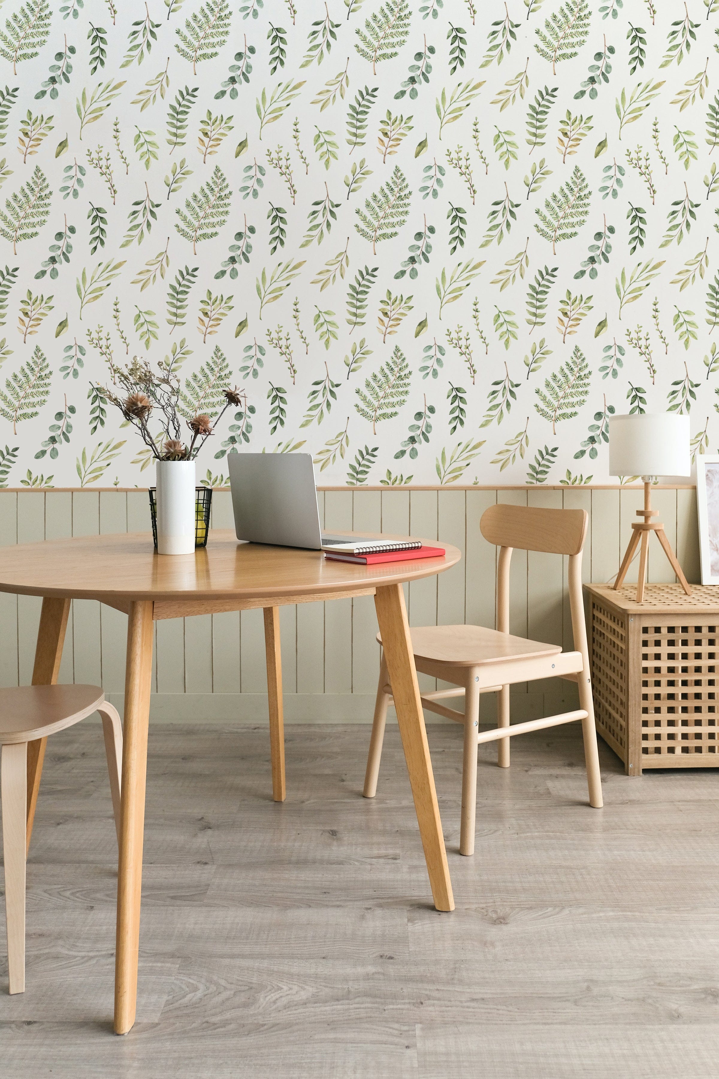 A cozy dining area with a wooden table and chairs, set against a wall decorated with Green Foliage Wallpaper, featuring hand-drawn green leaves and branches on a white background.