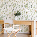 A stylish home office setup featuring Green Foliage Wallpaper with hand-drawn green leaves and branches on a white background, complemented by a wooden desk, a white chair, and a vase of greenery.