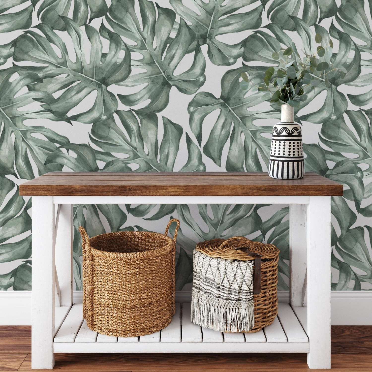 A stylish interior setting displaying the Modern Monstera Wallpaper as a feature wall behind a cozy white chair, accented by a striking black-and-white striped vase with greenery, evoking a modern jungle feel in the decor.