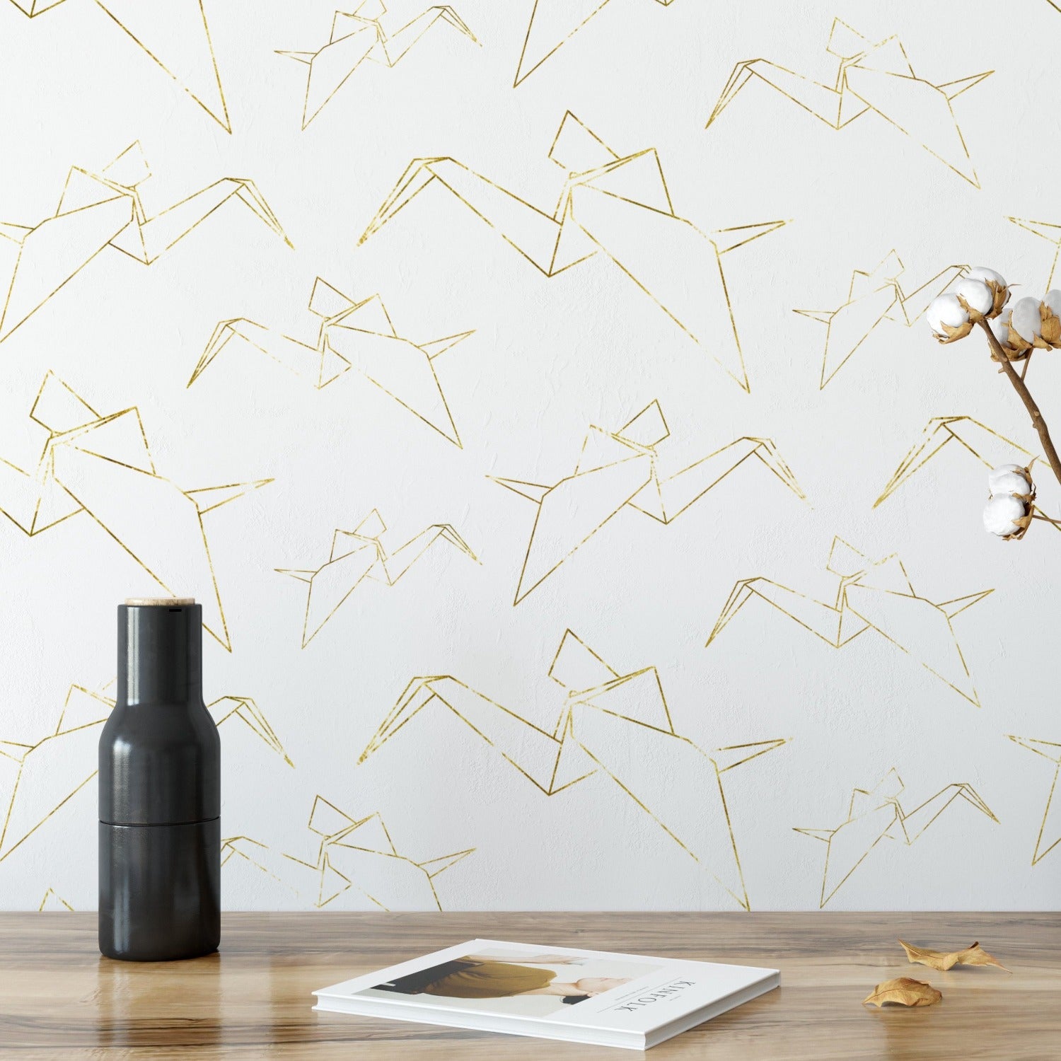 A room with white wallpaper adorned with gold origami crane patterns. The cranes are evenly spaced and create a harmonious design. A black vase and a cotton branch are placed on a wooden table in front of the wallpaper