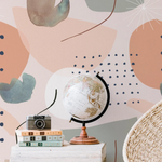 Interior scene with Modern Watercolour Abstract Wallpaper enhancing the room's decor. The wallpaper's large abstract watercolor shapes in pastel colors create a vibrant yet calming atmosphere, complemented by a wicker chair and vintage accessories