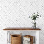 The 'Moroccan Tile Wallpaper VII - Steel Grey' elegantly adorns a wall above a simple white console table, complemented by woven baskets and a vase with greenery, enhancing the homey and stylish atmosphere