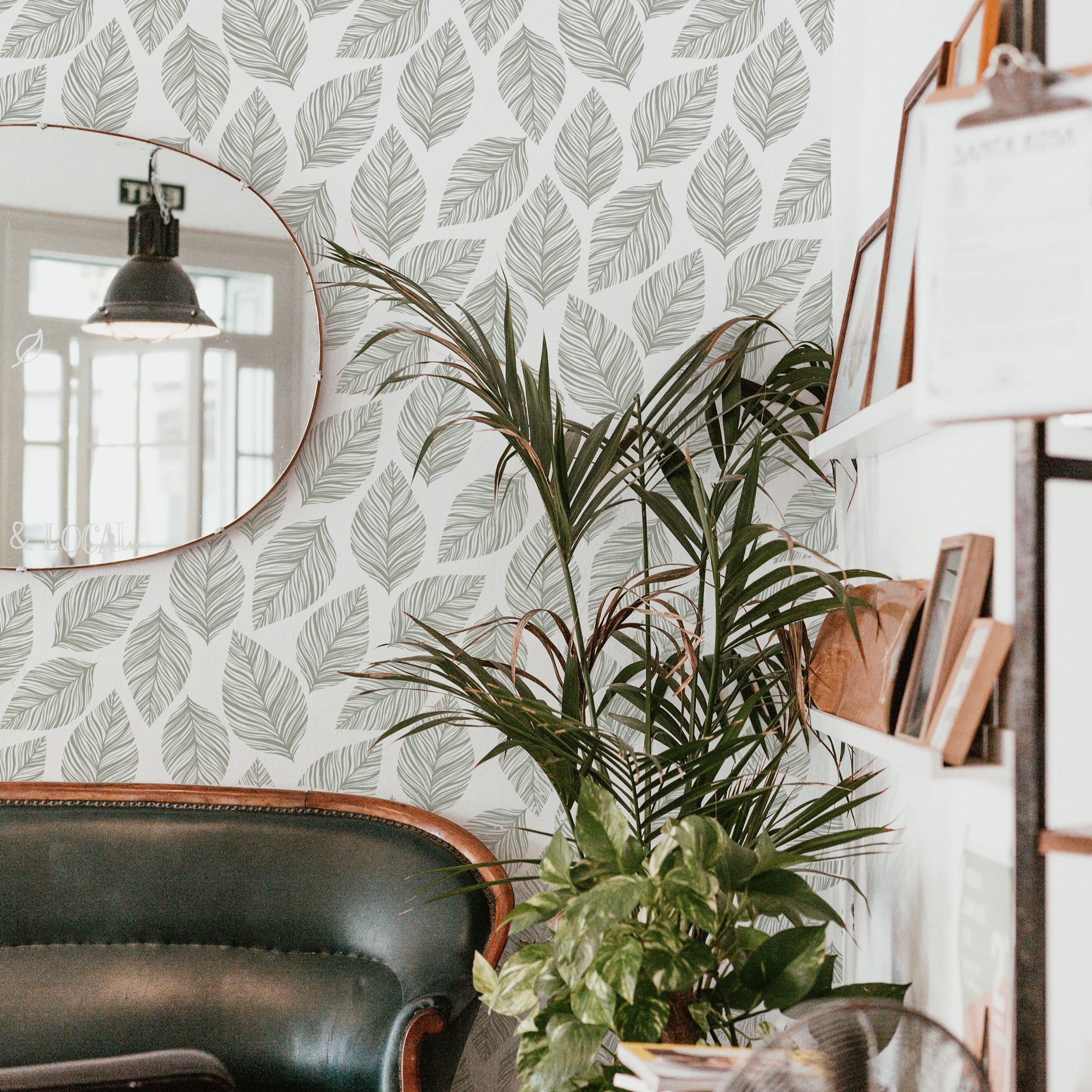  cozy corner of a cafe decorated with Leafy Wallpaper that showcases a clean and serene arrangement of leaf designs in grey on white. The natural theme is complemented by a vintage leather sofa and a vibrant potted plant, creating a welcoming atmosphere.