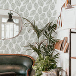 cozy corner of a cafe decorated with Leafy Wallpaper that showcases a clean and serene arrangement of leaf designs in grey on white. The natural theme is complemented by a vintage leather sofa and a vibrant potted plant, creating a welcoming atmosphere.