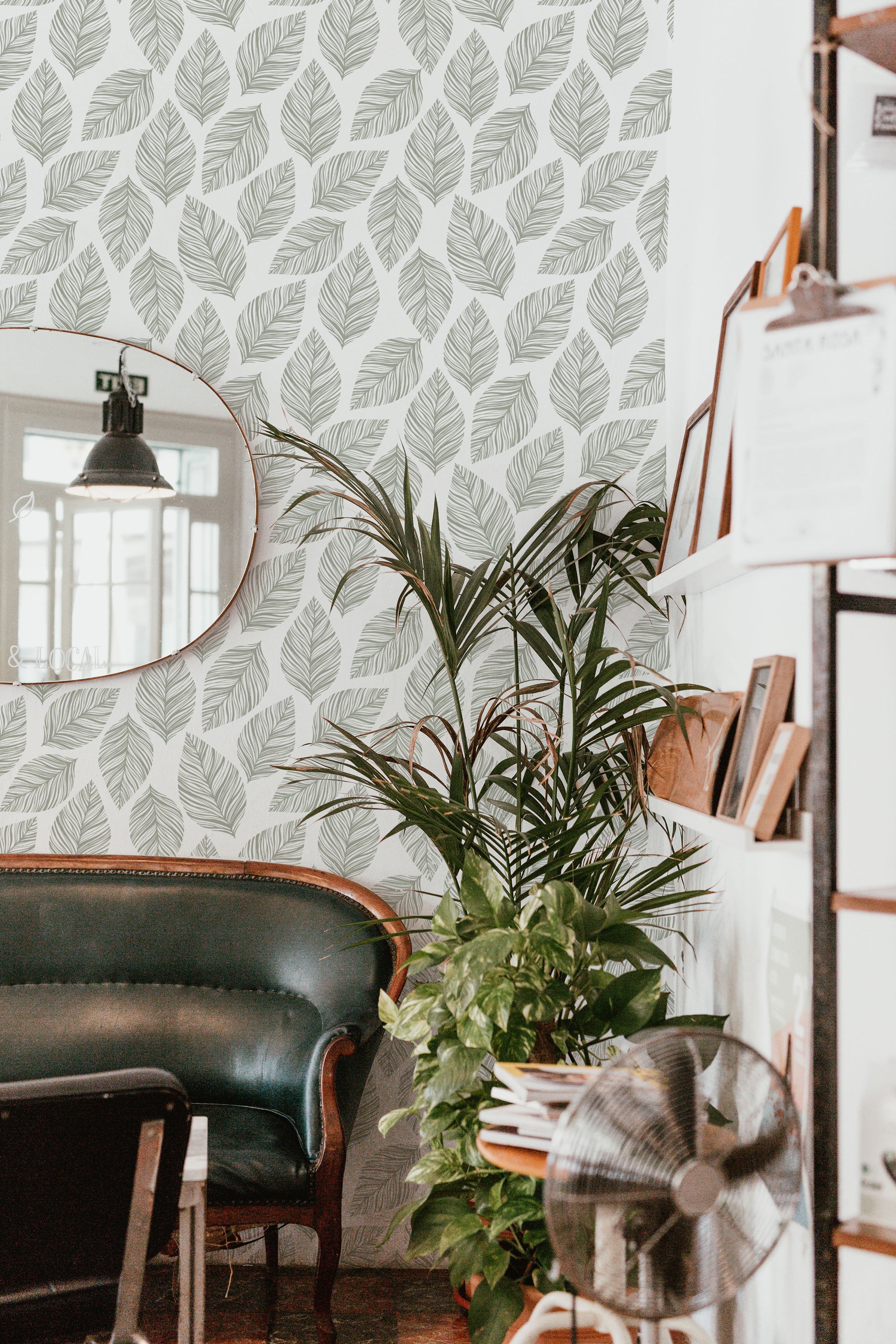 cozy corner of a cafe decorated with Leafy Wallpaper that showcases a clean and serene arrangement of leaf designs in grey on white. The natural theme is complemented by a vintage leather sofa and a vibrant potted plant, creating a welcoming atmosphere.