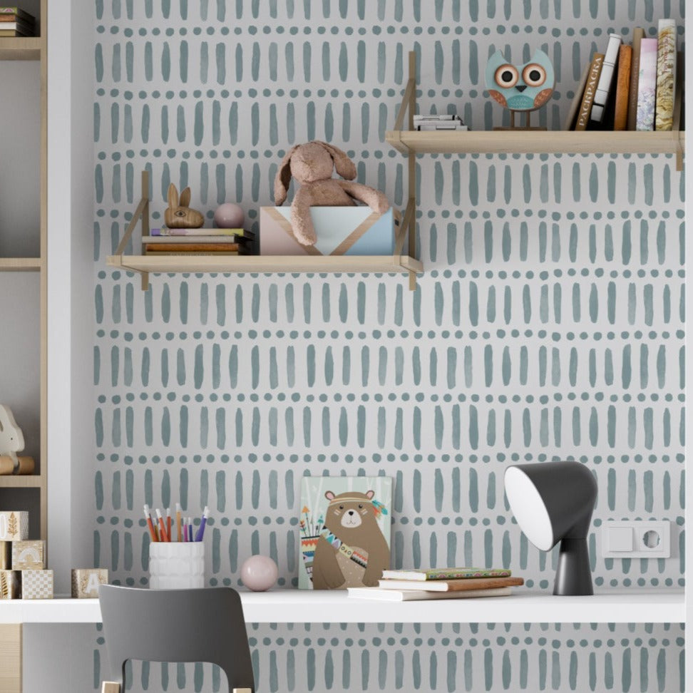 A child’s study area with walls covered in the Big Boho 44 Wallpaper in Blue Smoke. The playful yet soothing pattern of blue brush strokes and dots on a white backdrop provides a calming atmosphere, complementing the light wood furniture and whimsical shelf decorations.