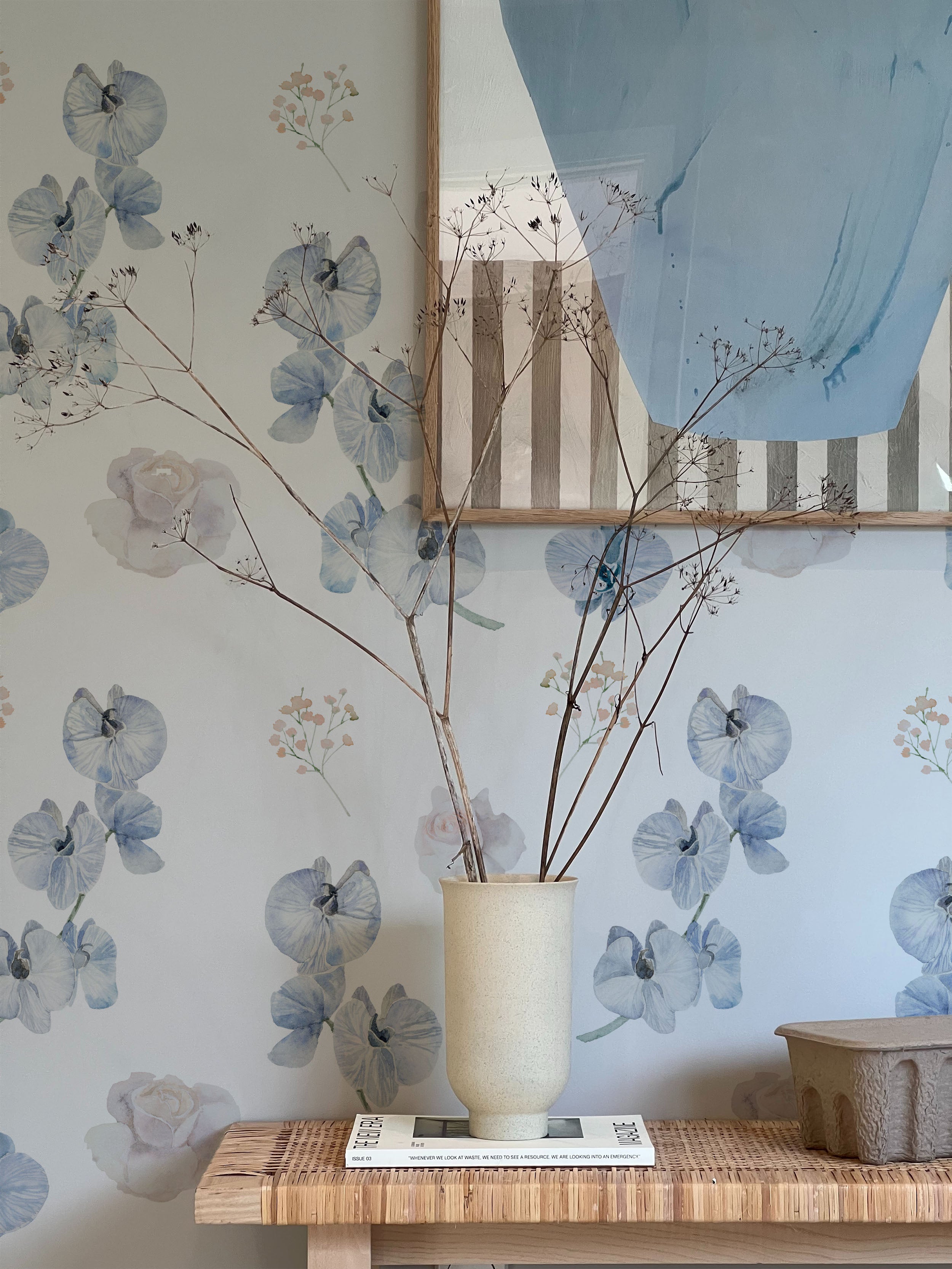 A stylish vignette with a vase of dried flowers on a wooden table against a wall covered in dreamy orchid wallpaper. The wallpaper features a delicate pattern of blue orchids, soft pink roses, and small sprigs of baby's breath on a light background, adding a touch of elegance and serenity to the space. An abstract painting hangs above the table.