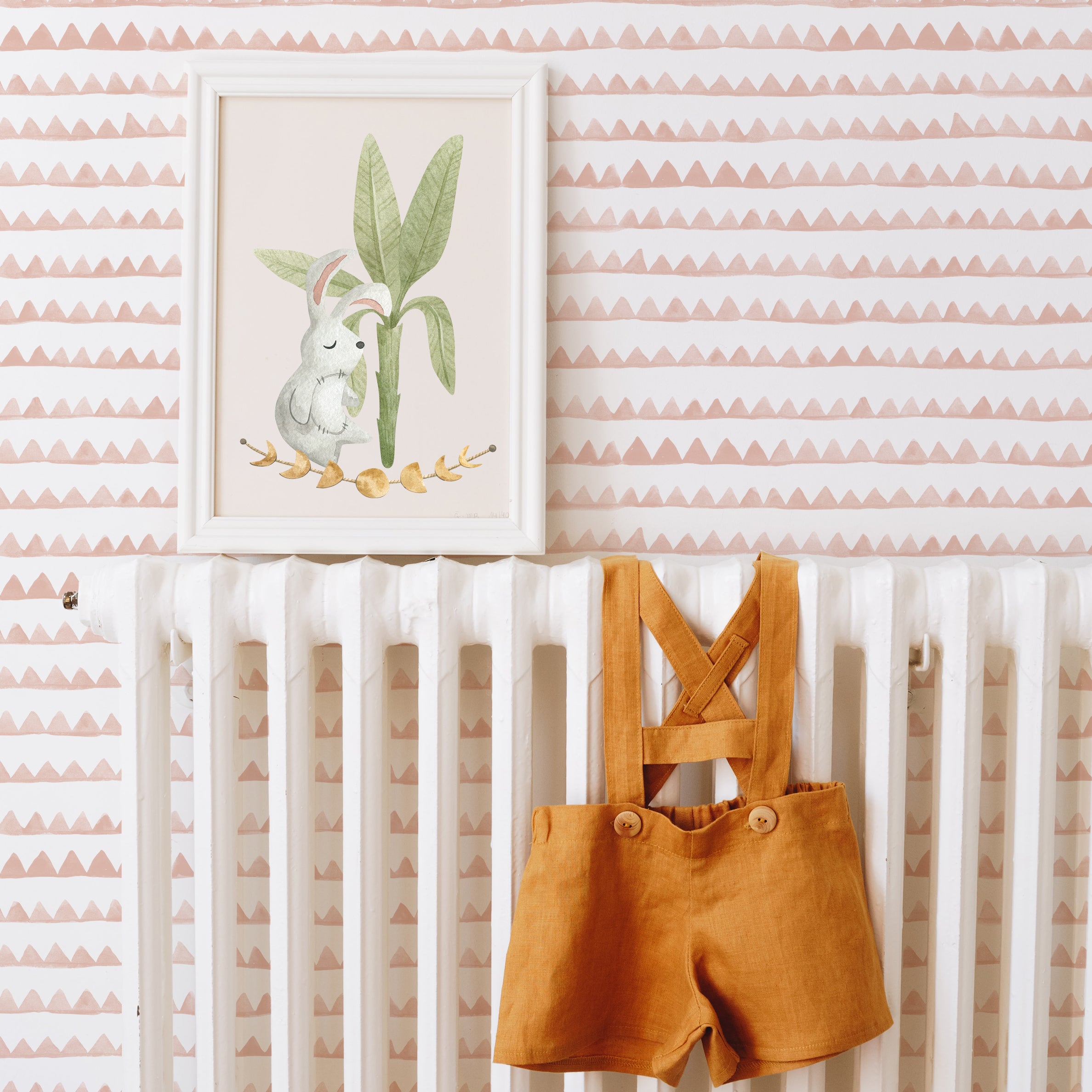 A charming nursery setup featuring the Kids Boho Wallpaper - 46, with muted terracotta zigzag patterns on a white background. The scene includes a framed illustration of a rabbit under a plant, and mustard-colored baby overalls hung on a white crib, creating a warm and inviting atmosphere.