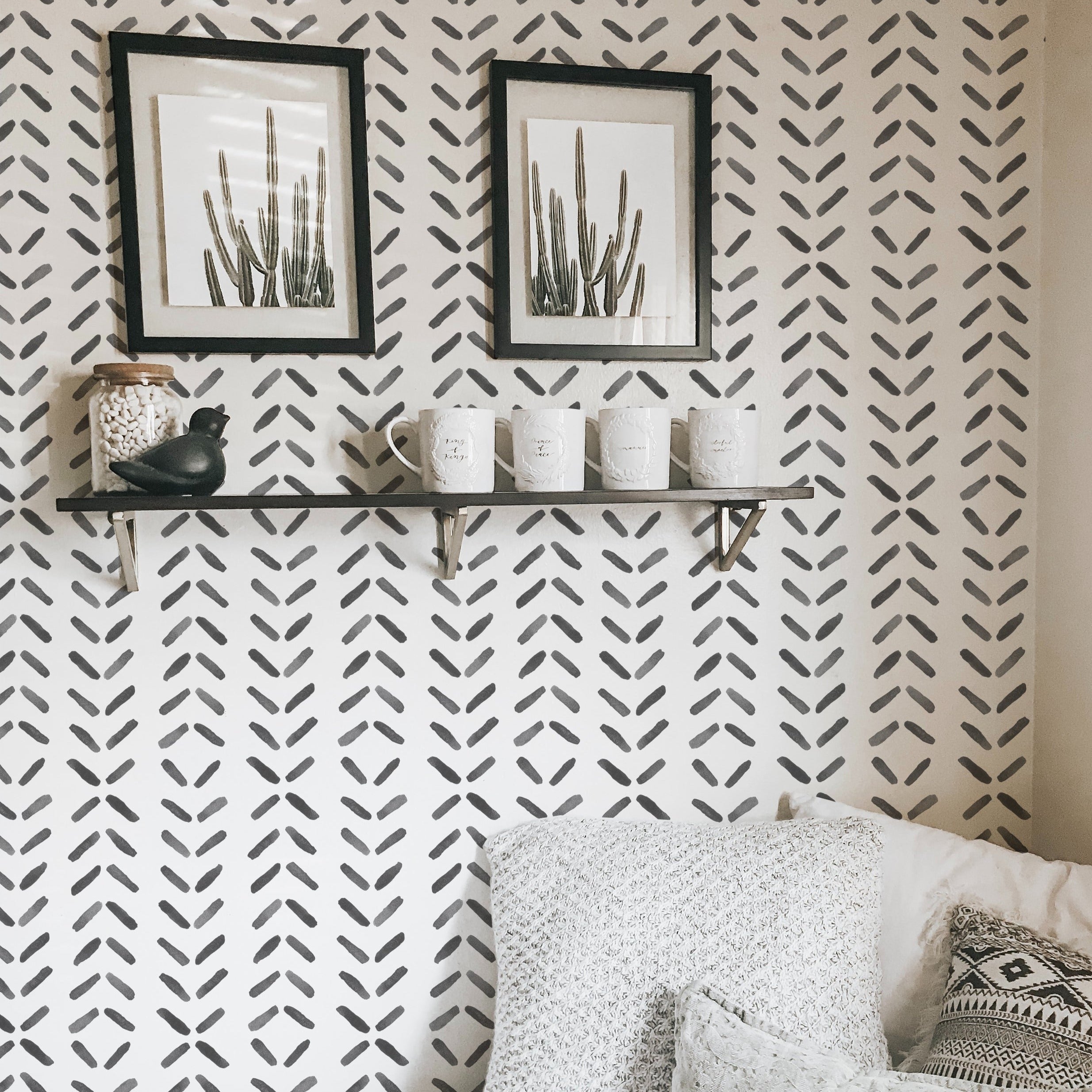 A cozy nook adorned with Boho Chevron Wallpaper featuring a repeating pattern of black chevron designs on a light background. This stylish setting is complemented by decorative shelves holding framed artwork and ceramic items, enhancing the room's modern bohemian aesthetic.