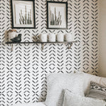 A cozy nook adorned with Boho Chevron Wallpaper featuring a repeating pattern of black chevron designs on a light background. This stylish setting is complemented by decorative shelves holding framed artwork and ceramic items, enhancing the room's modern bohemian aesthetic.