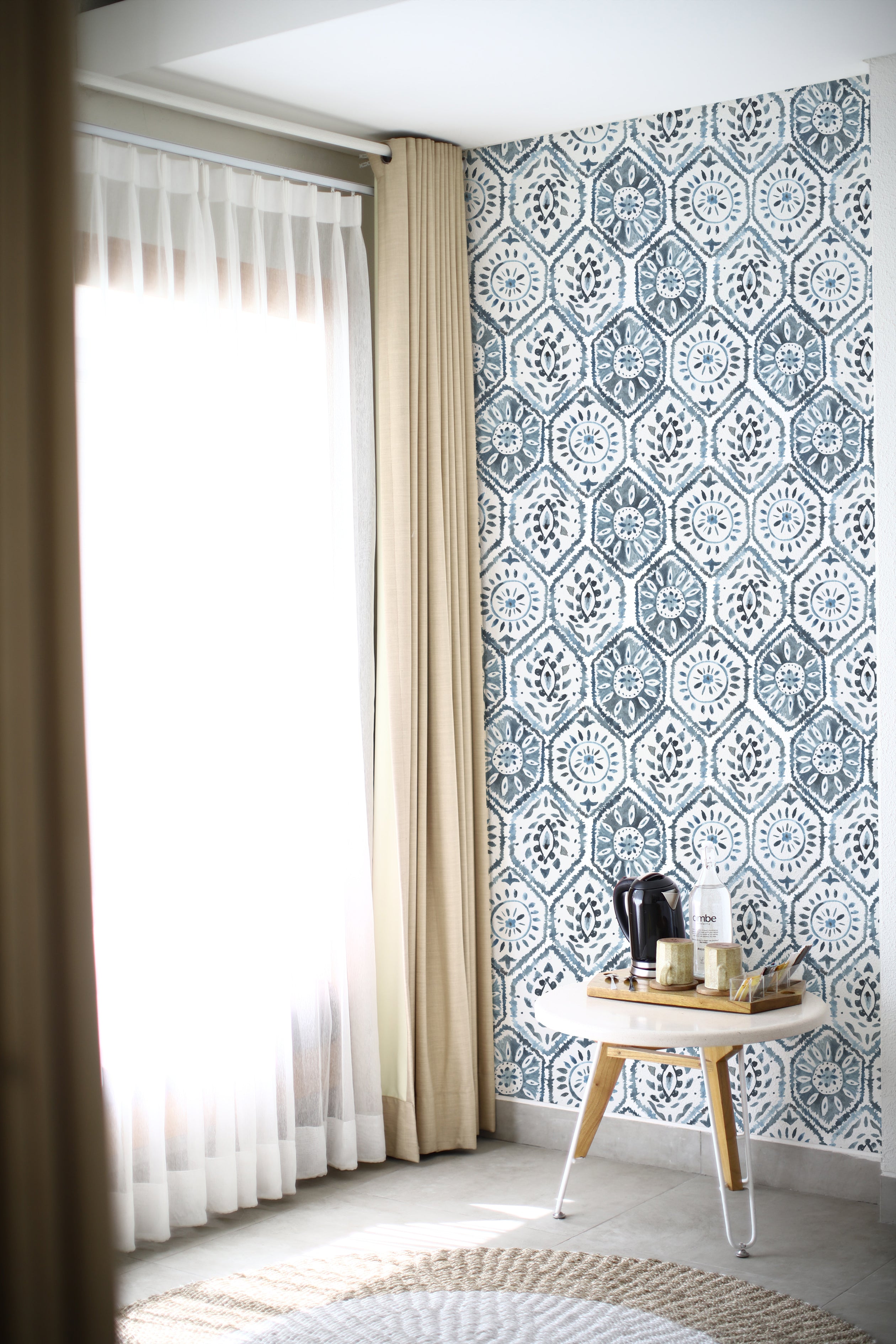 A cozy corner with sunlight filtering through sheer curtains next to a patterned Moroccan Tile wall in indigo blue, accented by a small wooden side table with a coffee maker and cups.