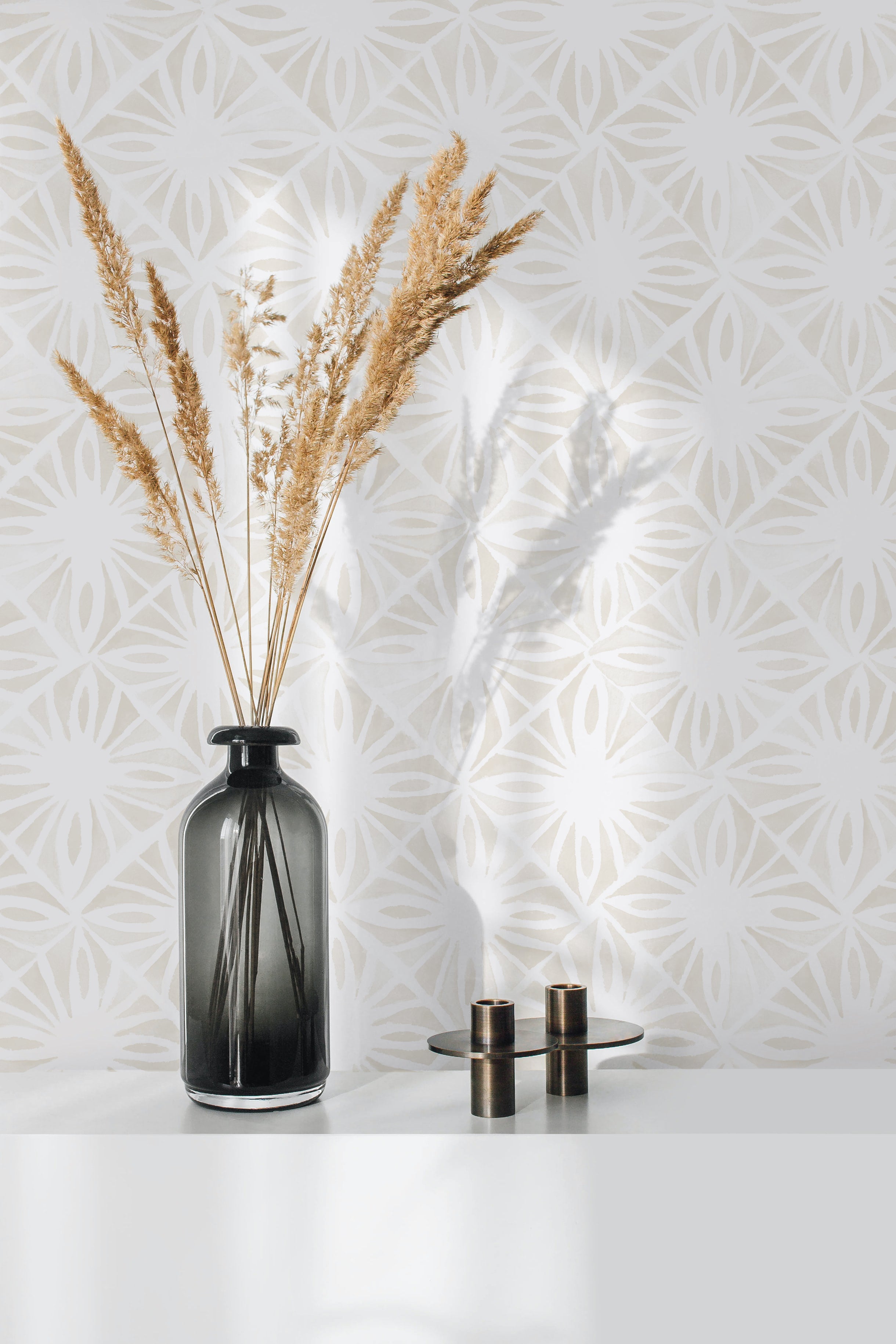 A minimalist home workspace is elevated by the Moroccan Tile Wallpaper II - Ecru, displaying an intricate pattern in soft ecru on a white background. The wallpaper adds a subtle, elegant touch to the room, complemented by a sleek black vase with dried pampas grass and simple metallic candle holders.