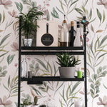 A modern shelving unit against a wall covered with the Botanic Floral Wallpaper, which is bursting with watercolor botanical illustrations. The shelves hold an assortment of houseplants, cookbooks, and bottles, enhancing the botanical theme in a functional space