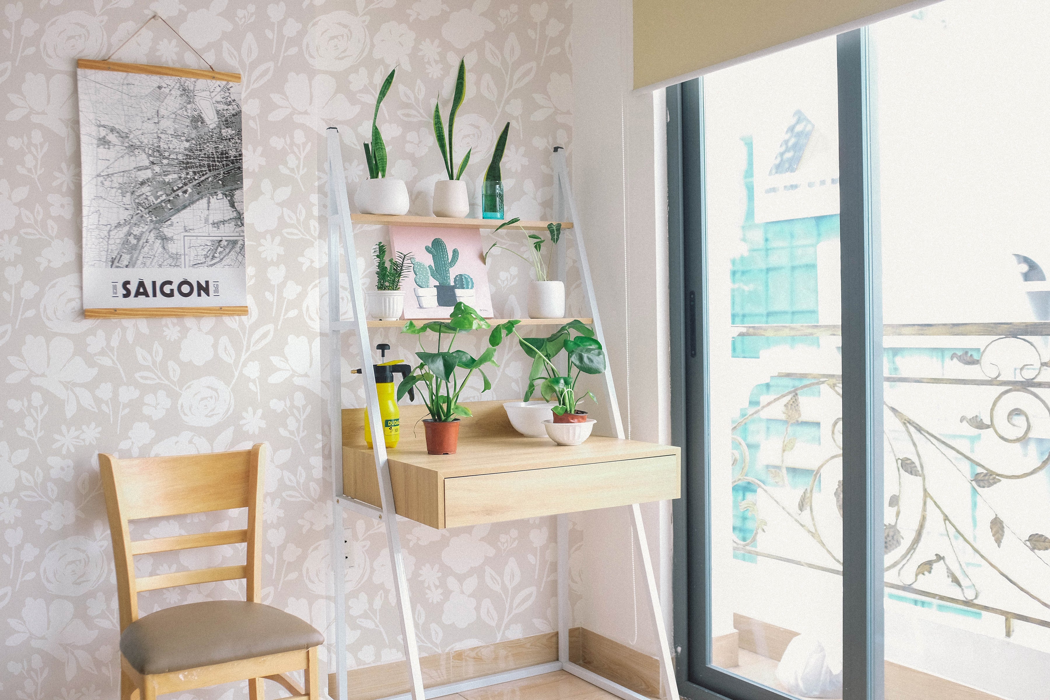 A cozy corner of a room featuring the Silhouette Floral Wallpaper in Faded Linen, adorned with a light wooden chair and a white shelf housing an array of potted green plants. A framed map of Saigon hangs on the wall, complementing the wallpaper's soft floral design.