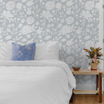 A cozy bedroom interior showcasing walls adorned with pale blue floral silhouette wallpaper. The decor includes a white bed with blue pillows, complemented by a wooden bedside table and a potted plant, enhancing the room’s serene and fresh atmosphere.