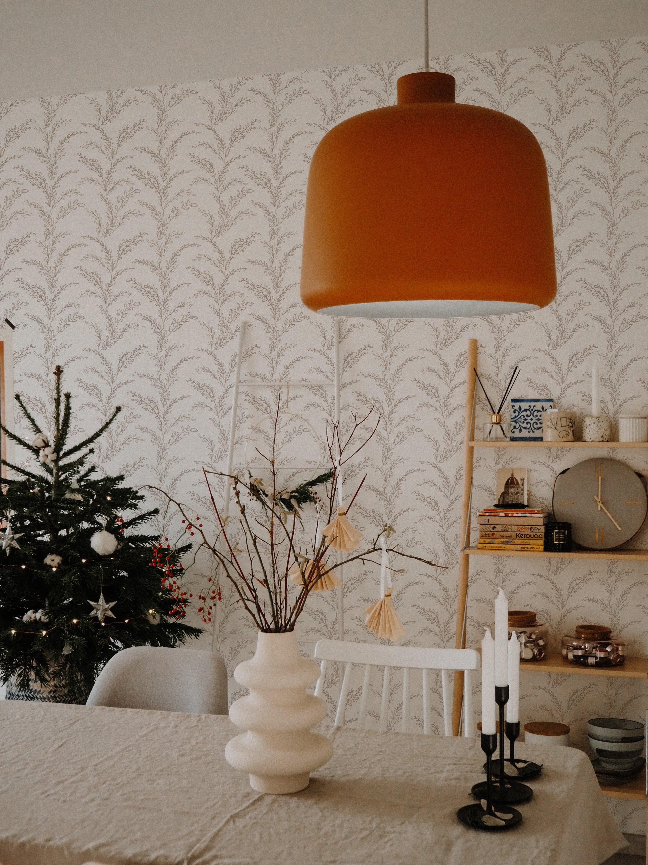 A cozy dining area adorned with the Rustic Foliage Wallpaper in Beige, featuring an intricate botanical print. The space is warmly lit by an oversized orange pendant lamp above a white table with modern gray and white chairs. The corner includes a tastefully decorated Christmas tree and a striking white sculptural vase with bare branches.