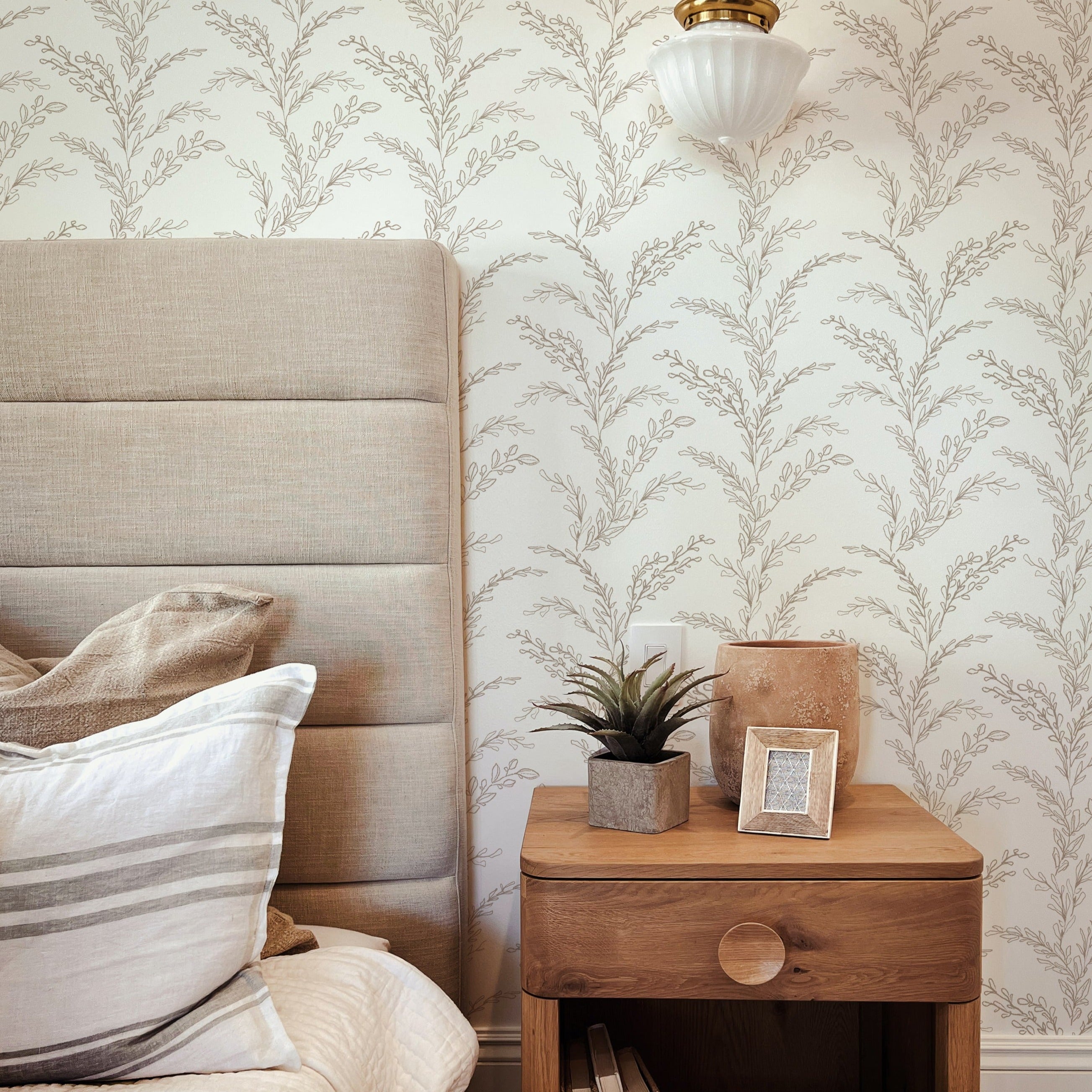 An elegant bedroom showcasing the Rustic Foliage Wallpaper in Beige, providing a serene backdrop to a fabric headboard and a wooden bedside table with a potted plant, a beige textured vase, and a small photo frame