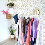 Spring Fling Wallpaper adorning a bright and airy room with a clothing rack displaying various colorful outfits, a potted snake plant, hanging basket with pink flowers, and a hat hanging on the rack