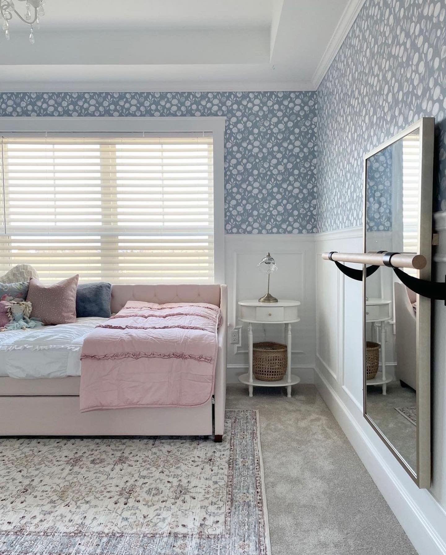 A light and airy bedroom with 'Subtle Botanica II - Inverted' wallpaper featuring a soothing pattern of white botanical silhouettes against a soft blue-grey background. The room is elegantly styled with a blush pink upholstered bed, a white bedside table, and a patterned rug, enhancing the serene aesthetic.