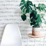 A minimalist interior scene showcasing the "Hand Painted Wallpaper" with a gentle sage green herringbone pattern. A modern white chair sits in the foreground, beside a small wooden table holding a potted monstera plant, which adds a vibrant touch of greenery against the hand-painted strokes of the wallpaper.
