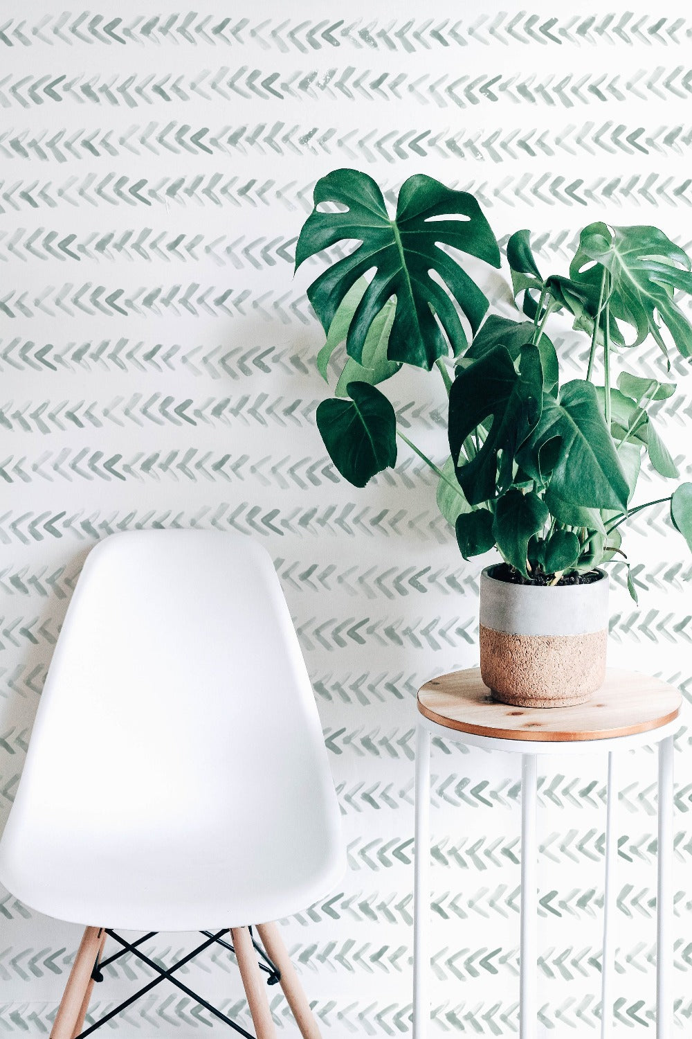 A minimalist interior scene showcasing the "Hand Painted Wallpaper" with a gentle sage green herringbone pattern. A modern white chair sits in the foreground, beside a small wooden table holding a potted monstera plant, which adds a vibrant touch of greenery against the hand-painted strokes of the wallpaper.