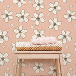 A cozy corner showcasing the Retro Pink Flowers Wallpaper, which enhances the space with its large white floral designs on a pink background, accompanied by a wooden stool topped with crocheted items.