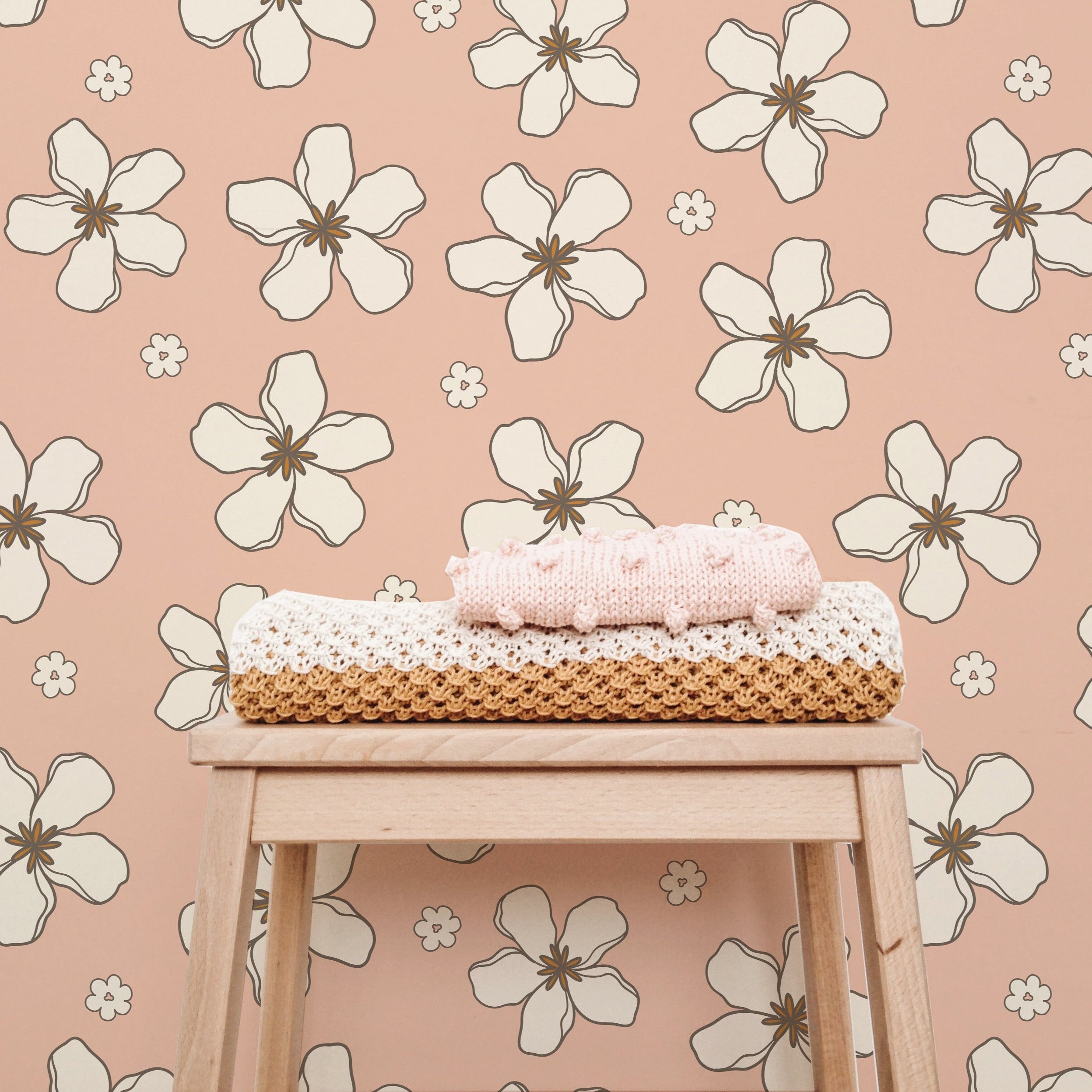 A cozy corner showcasing the Retro Pink Flowers Wallpaper, which enhances the space with its large white floral designs on a pink background, accompanied by a wooden stool topped with crocheted items.