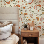 An elegant bedroom corner showcasing a tall, fabric-upholstered headboard against a vibrant Retro Pattern Wallpaper with a floral design featuring orange and pink blooms intertwined with green foliage. A wooden bedside table with a potted succulent, a square terracotta vase, and a small photo frame sits adjacent to the bed