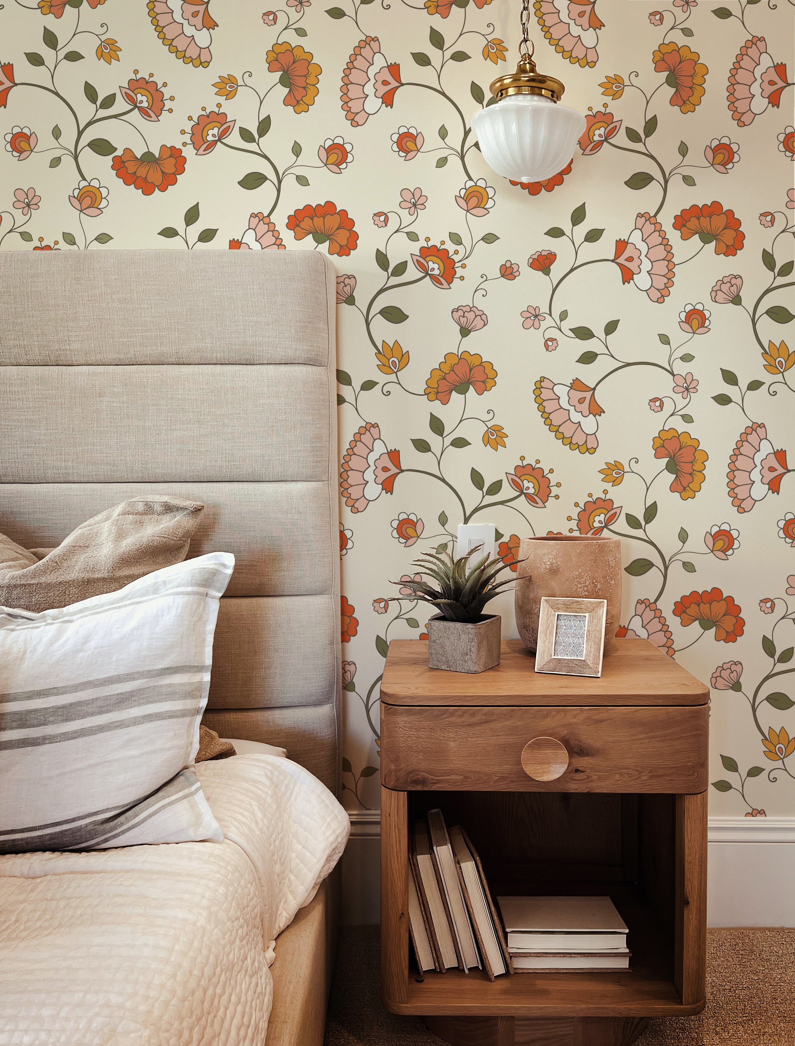 An elegant bedroom corner showcasing a tall, fabric-upholstered headboard against a vibrant Retro Pattern Wallpaper with a floral design featuring orange and pink blooms intertwined with green foliage. A wooden bedside table with a potted succulent, a square terracotta vase, and a small photo frame sits adjacent to the bed