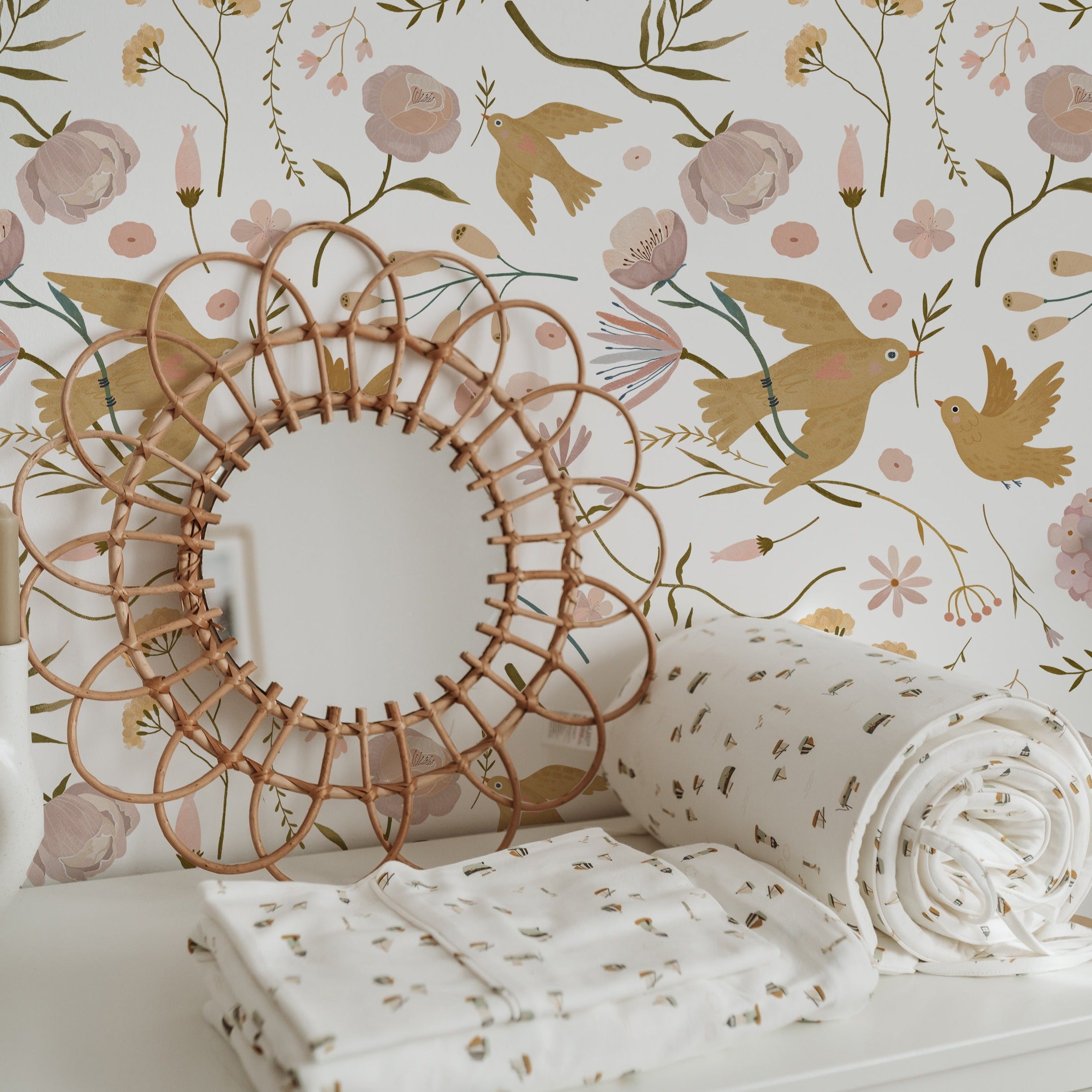 A decorative setting featuring the Pastel Garden Wallpaper, with a woven circular mirror and floral-printed textiles enhancing the wallpaper's intricate pattern of birds and blossoms.