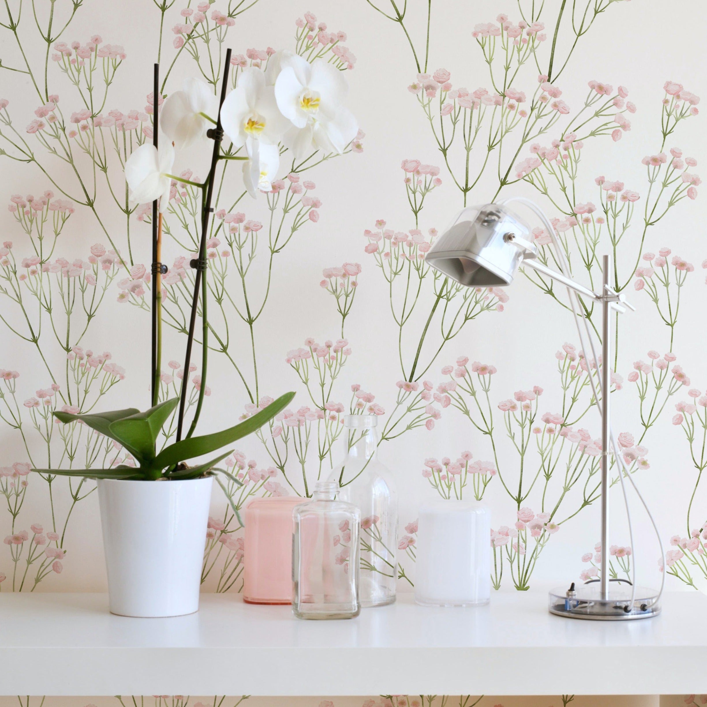 A cozy corner of a room enhanced by the light and airy 'Le Marchand de Fleurs Wallpaper', with its intricate pattern of green stems and pink blossoms. The wallpaper adds a romantic, garden-inspired atmosphere, complemented by an elegant white orchid and simple beauty accessories on a white shelf.