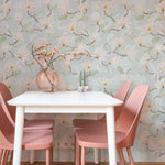 A cozy dining area with walls covered in 'Watercolour Magnolia Wallpaper II', creating a tranquil and inviting environment. A simple white table is flanked by dusty pink chairs, and a round textured vase with branches sits in the middle, echoing the wallpaper's natural motif.