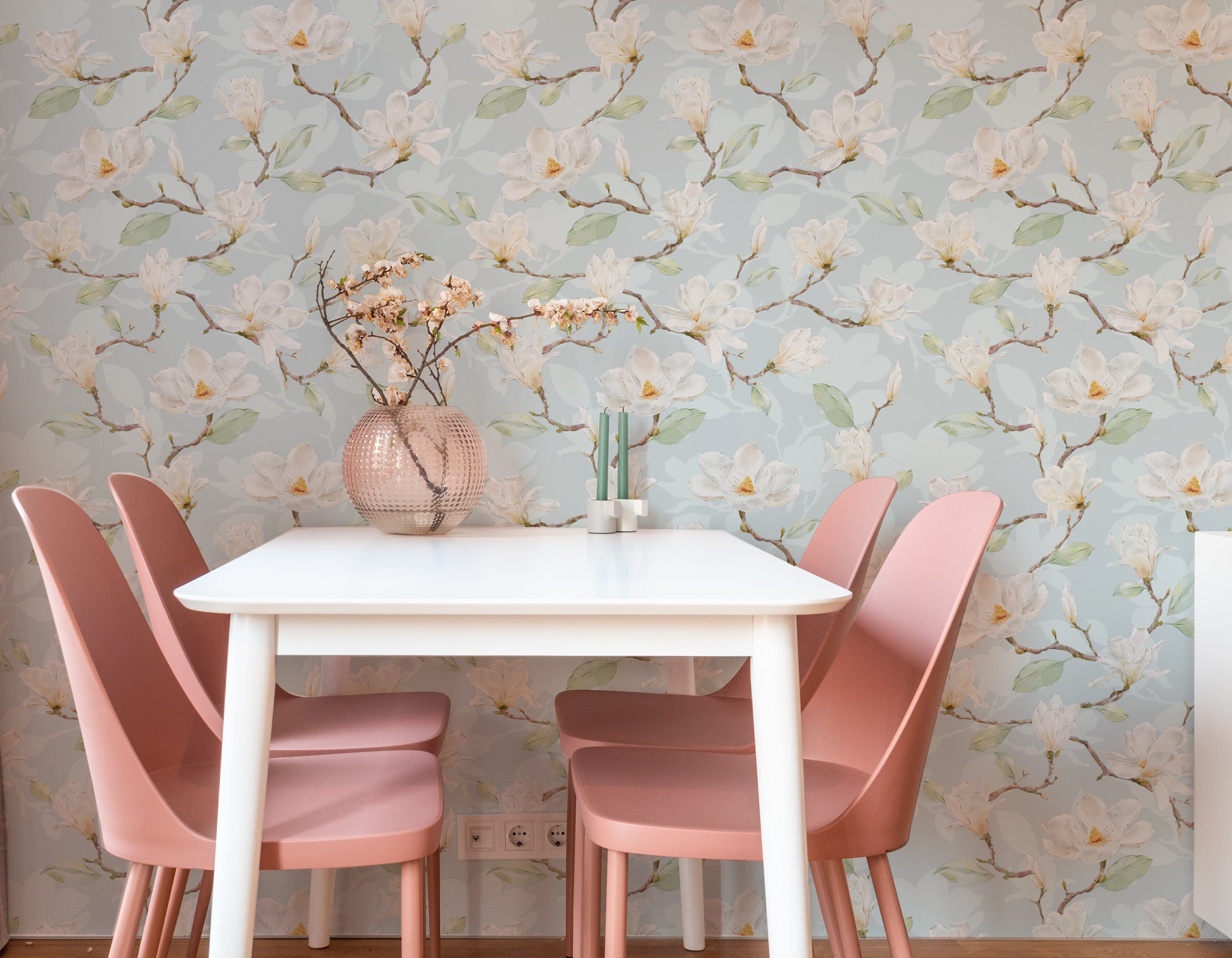 A cozy dining area with walls covered in 'Watercolour Magnolia Wallpaper II', creating a tranquil and inviting environment. A simple white table is flanked by dusty pink chairs, and a round textured vase with branches sits in the middle, echoing the wallpaper's natural motif.