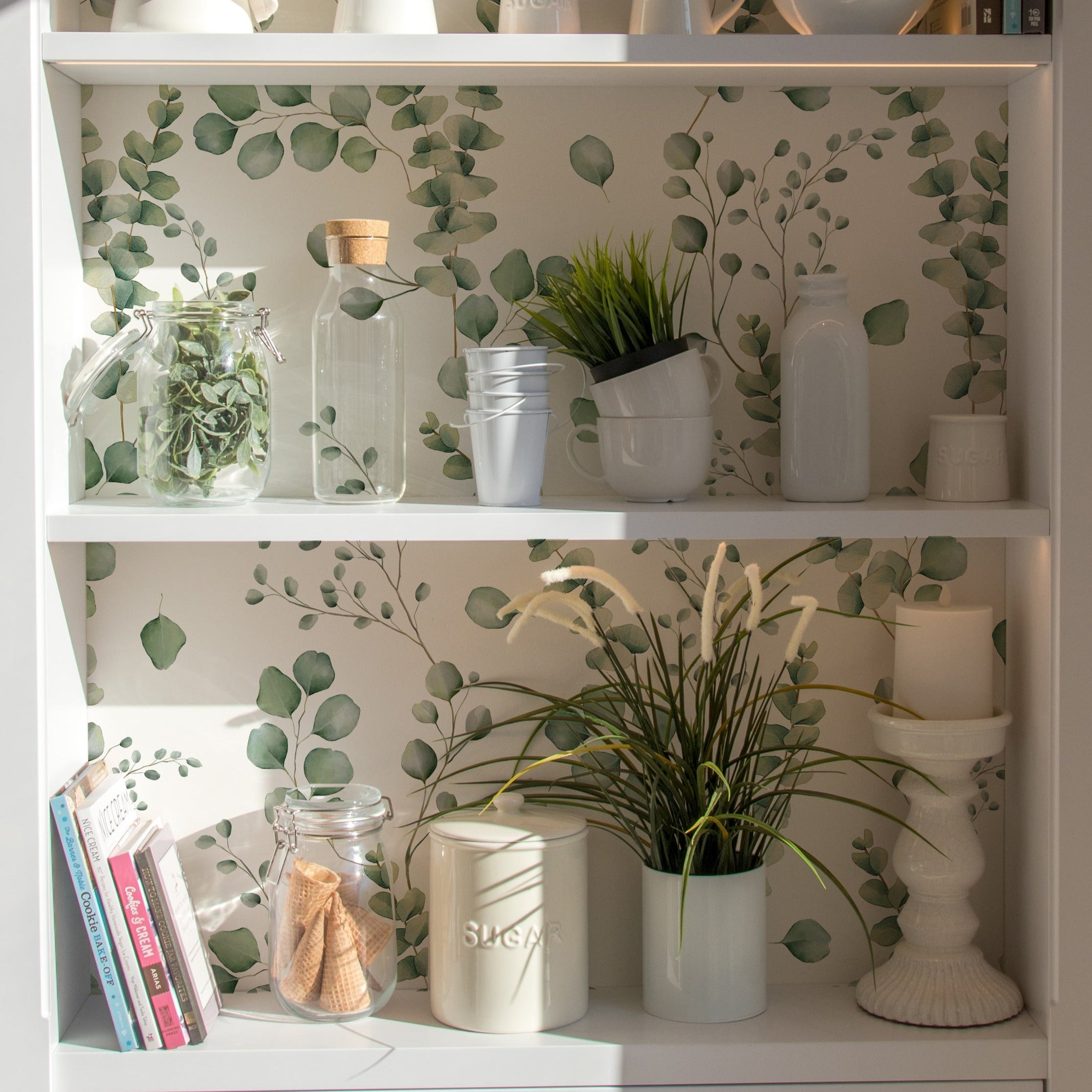 A styled interior setting showcasing shelves lined with the same eucalyptus leaf wallpaper. The shelves are decorated with items like books, white ceramic jars, and green plants, creating a cohesive, tranquil decor theme that complements the wallpaper's botanical design.