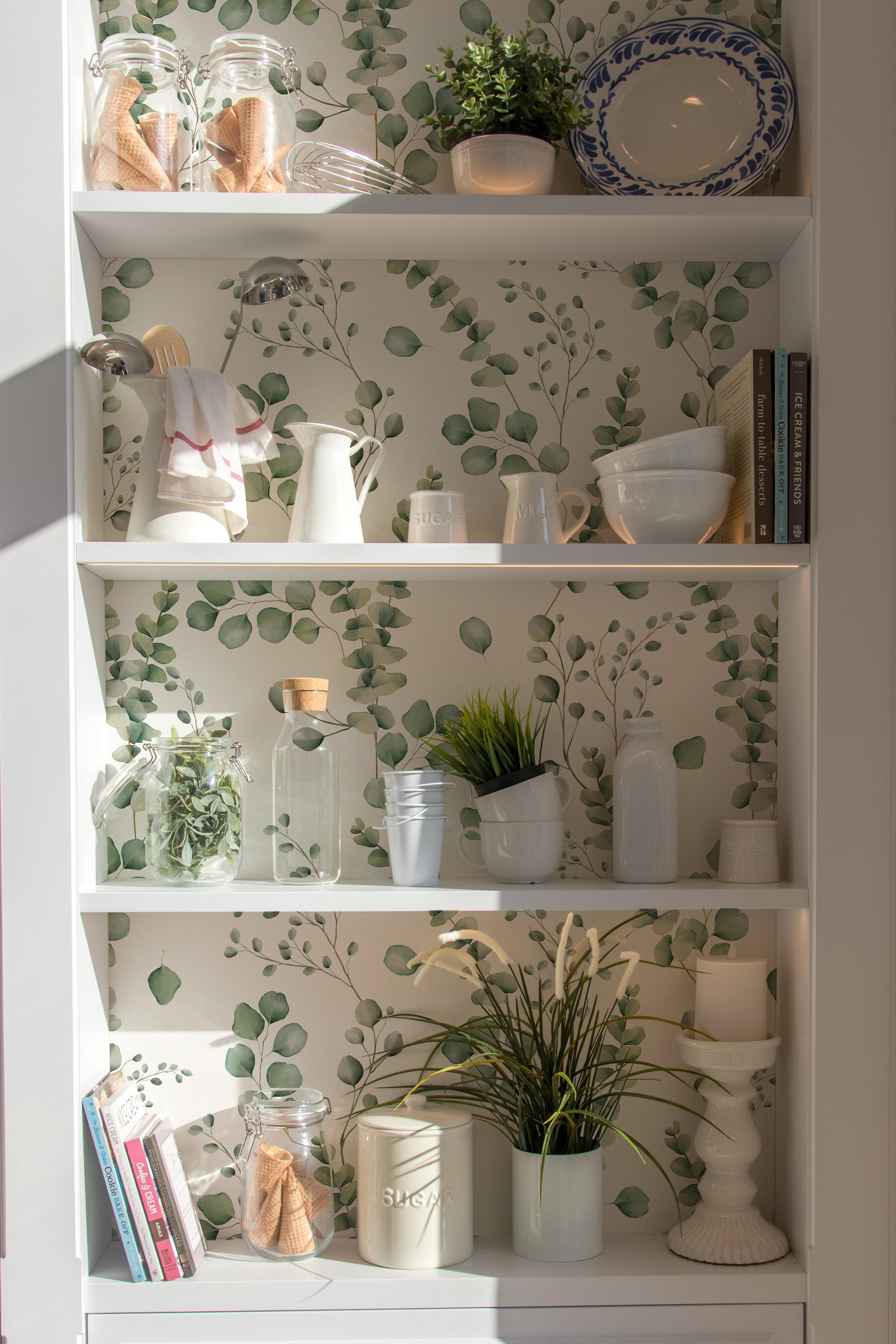 A styled interior setting showcasing shelves lined with the same eucalyptus leaf wallpaper. The shelves are decorated with items like books, white ceramic jars, and green plants, creating a cohesive, tranquil decor theme that complements the wallpaper's botanical design.