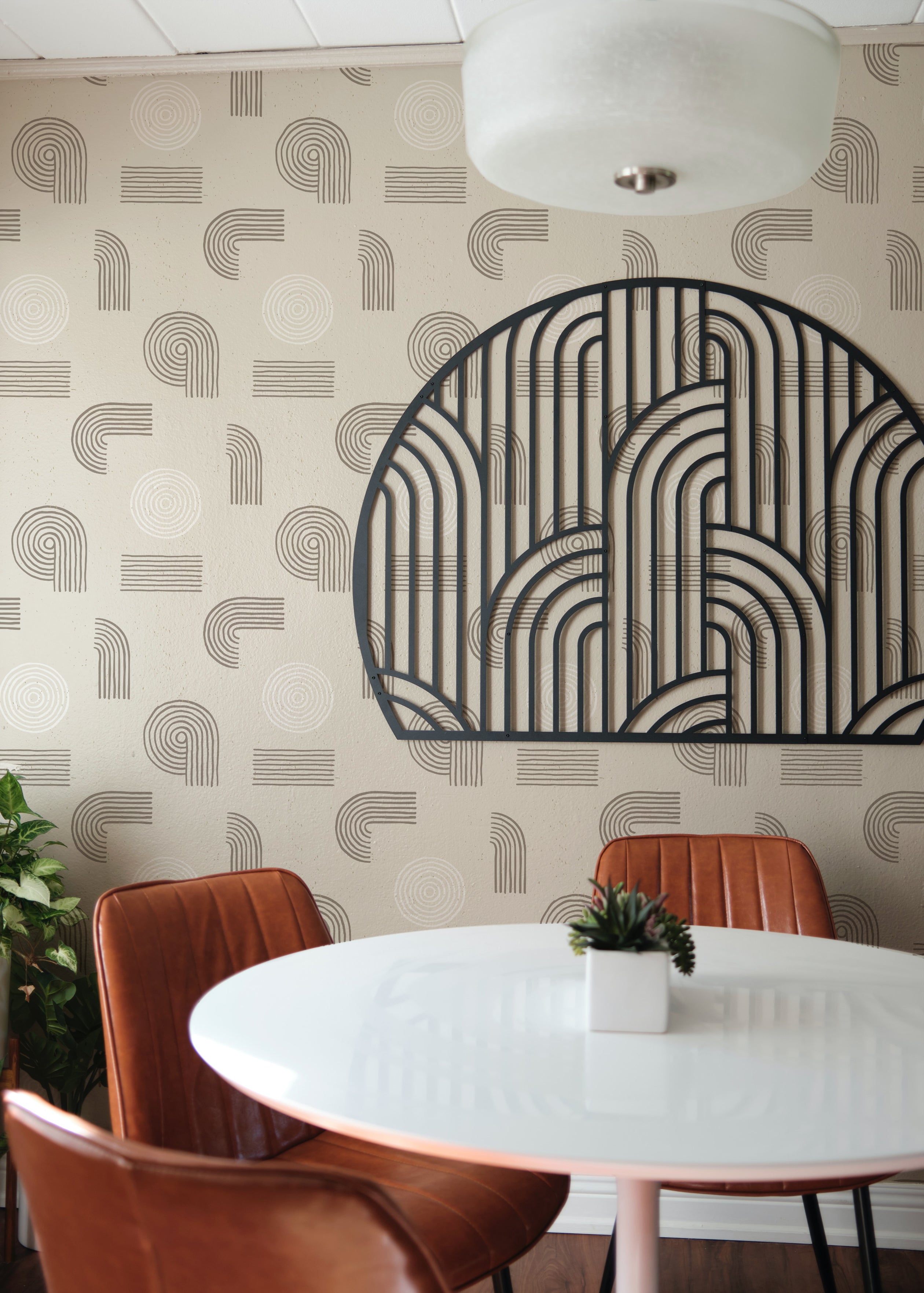 A modern dining space featuring Sand Zen Geometric Wallpaper - VII, characterized by abstract patterns of circles and curved lines in muted beige and off-white tones. The space includes a white round table, brown leather chairs, and a decorative metal wall art, enhancing the minimalist aesthetic.