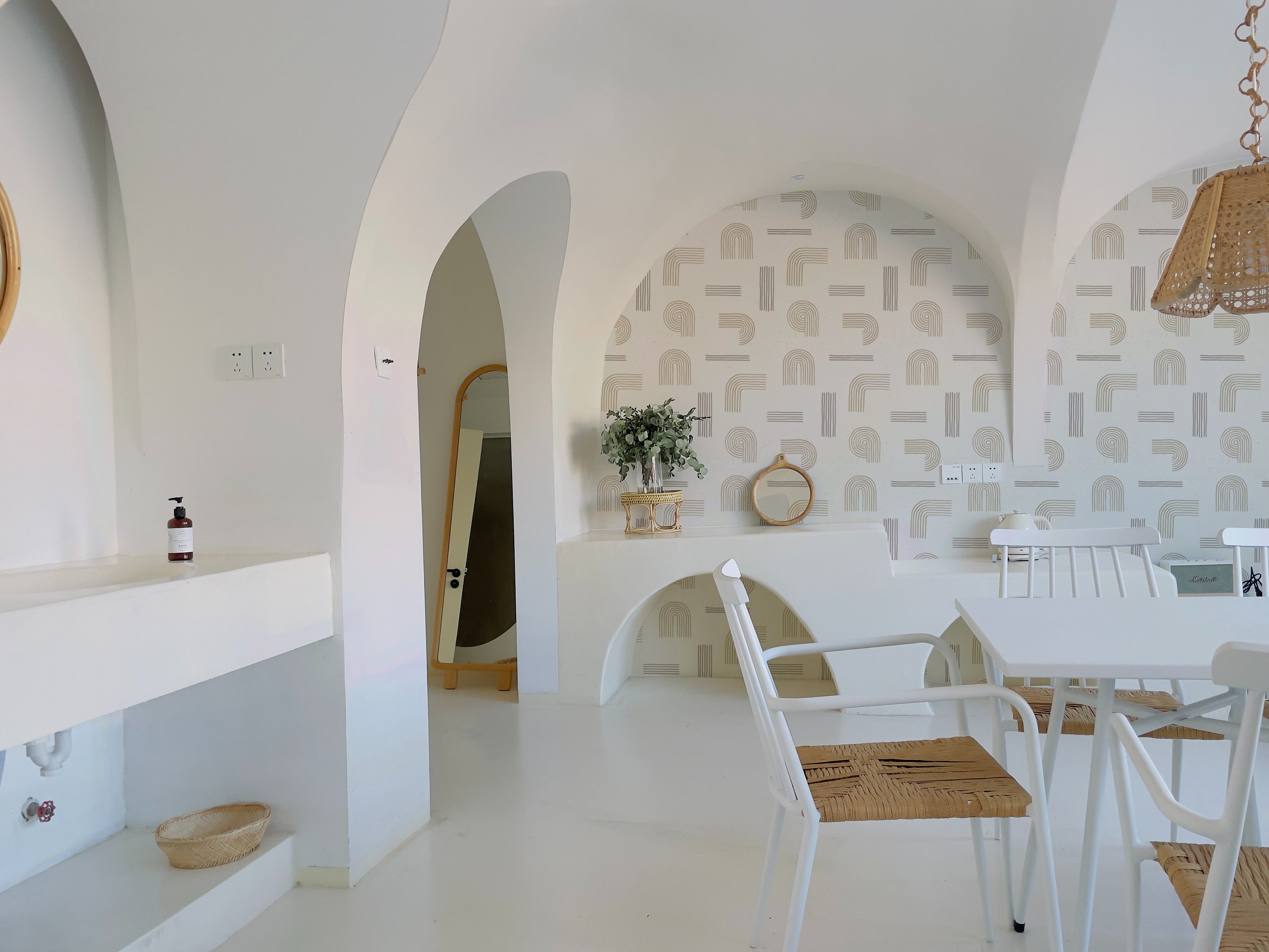 A bright and airy room decorated with Sand Zen Geometric Wallpaper - VII on the walls, complemented by white furnishings and natural wood accents. The space is styled with minimalist decor, including a white arch-shaped mirror and a small plant, creating a serene atmosphere.