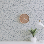 A well-composed interior scene using the "Subtle Botanica - Pale Blue" wallpaper to create a peaceful and refreshing wall accent. The pale blue leaf patterns contribute to a tranquil atmosphere, complemented by a wooden clock, a modern white desk lamp, and a potted plant. The wallpaper's design successfully creates an organic and calming environment ideal for a study room or living space.