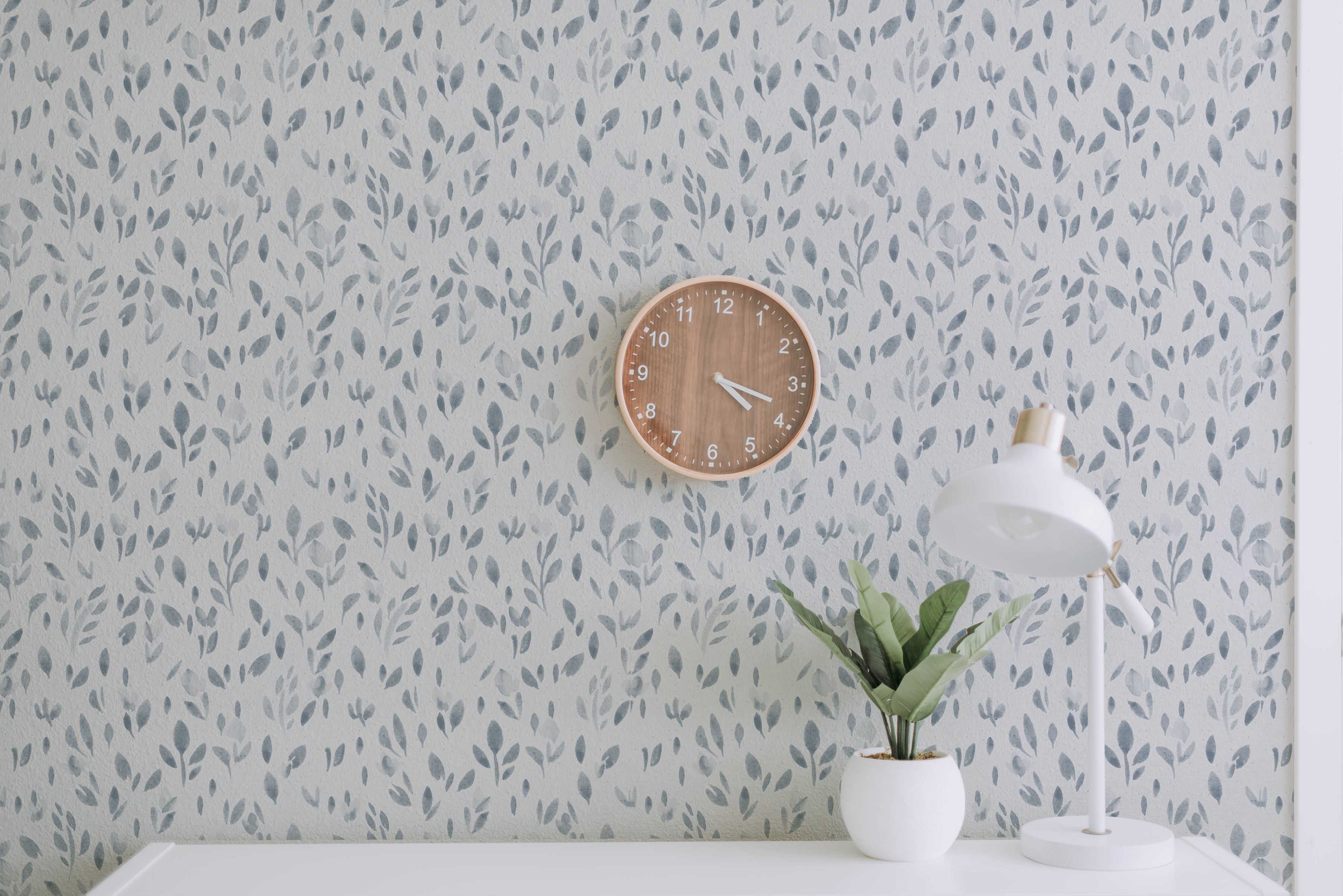 A well-composed interior scene using the "Subtle Botanica - Pale Blue" wallpaper to create a peaceful and refreshing wall accent. The pale blue leaf patterns contribute to a tranquil atmosphere, complemented by a wooden clock, a modern white desk lamp, and a potted plant. The wallpaper's design successfully creates an organic and calming environment ideal for a study room or living space.