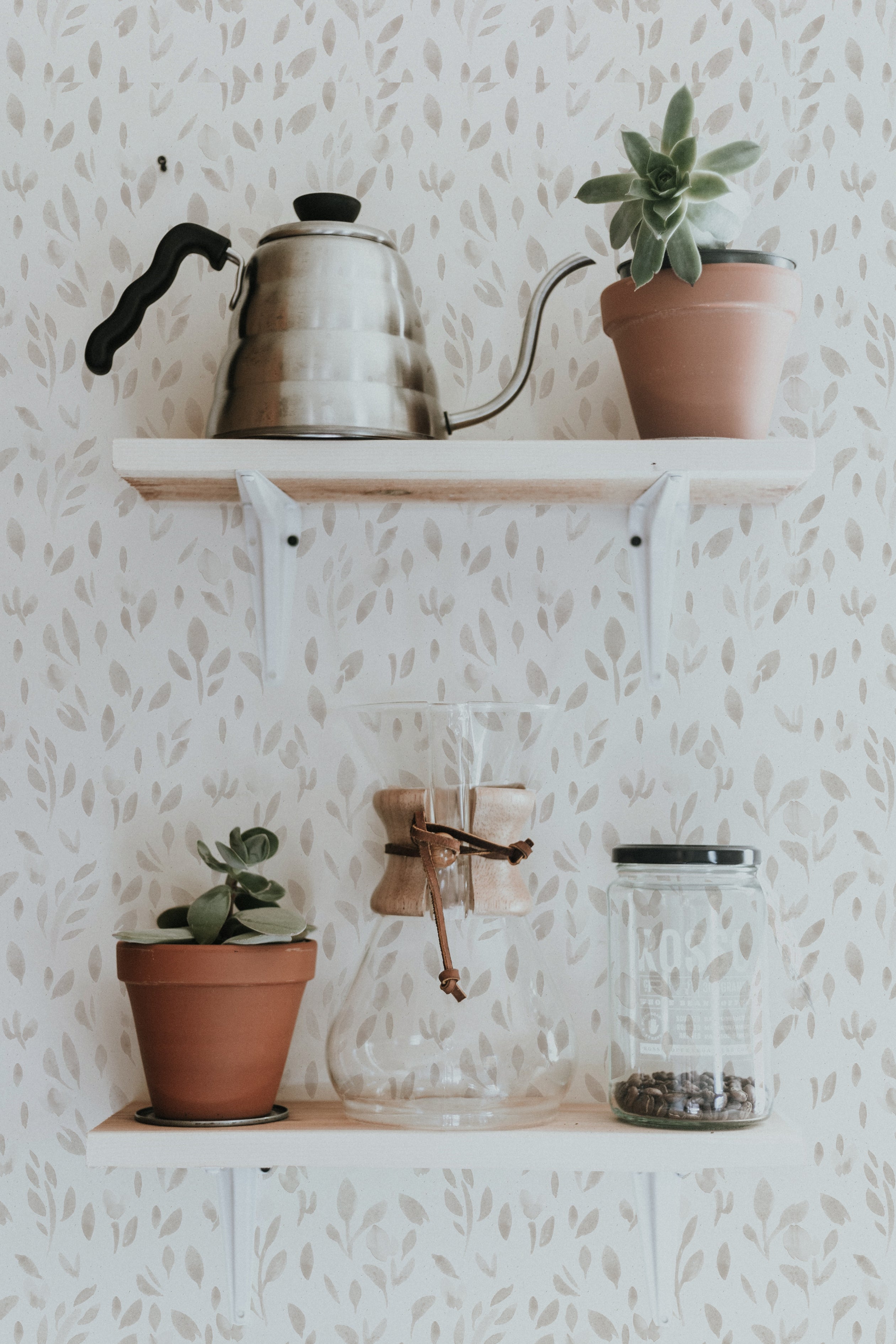 A homey kitchen scene with 'Subtle Botanica - Linen' wallpaper, displaying a pattern of soft gray leaves on a light linen background. Two white floating shelves hold a classic metal kettle, terracotta pots with green succulents, and glass jars.