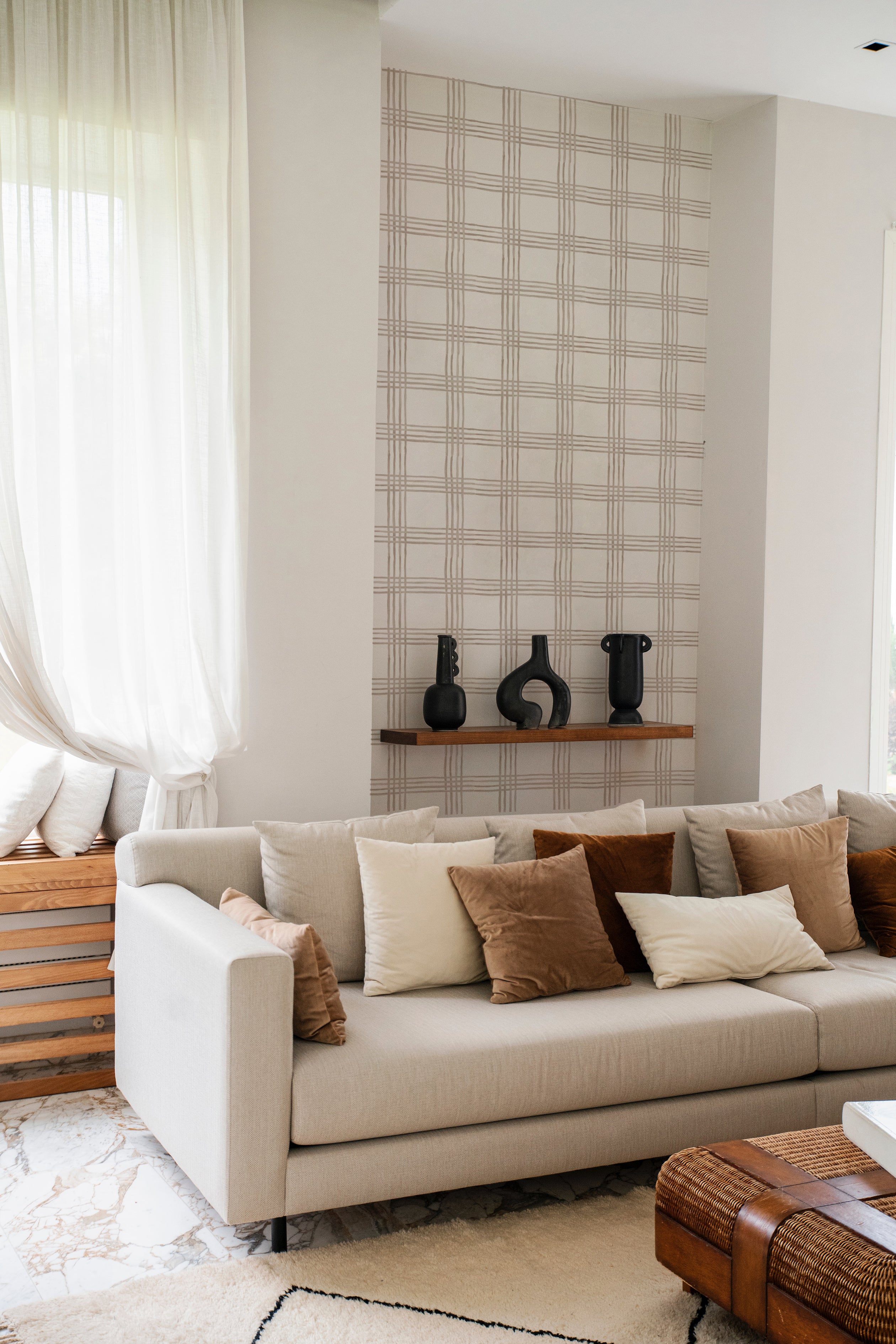 An elegant living room scene featuring the Big Boho Wallpaper - 123 on one wall. The wallpaper’s grid pattern in beige on a white background complements a cozy sofa filled with pillows in earthy tones and a wooden shelf displaying unique black vases, enhancing the room's modern aesthetic