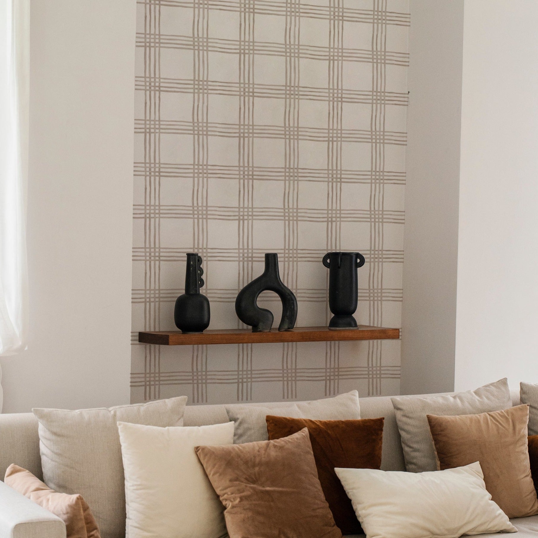 An elegant living room scene featuring the Big Boho Wallpaper - 123 on one wall. The wallpaper’s grid pattern in beige on a white background complements a cozy sofa filled with pillows in earthy tones and a wooden shelf displaying unique black vases, enhancing the room's modern aesthetic